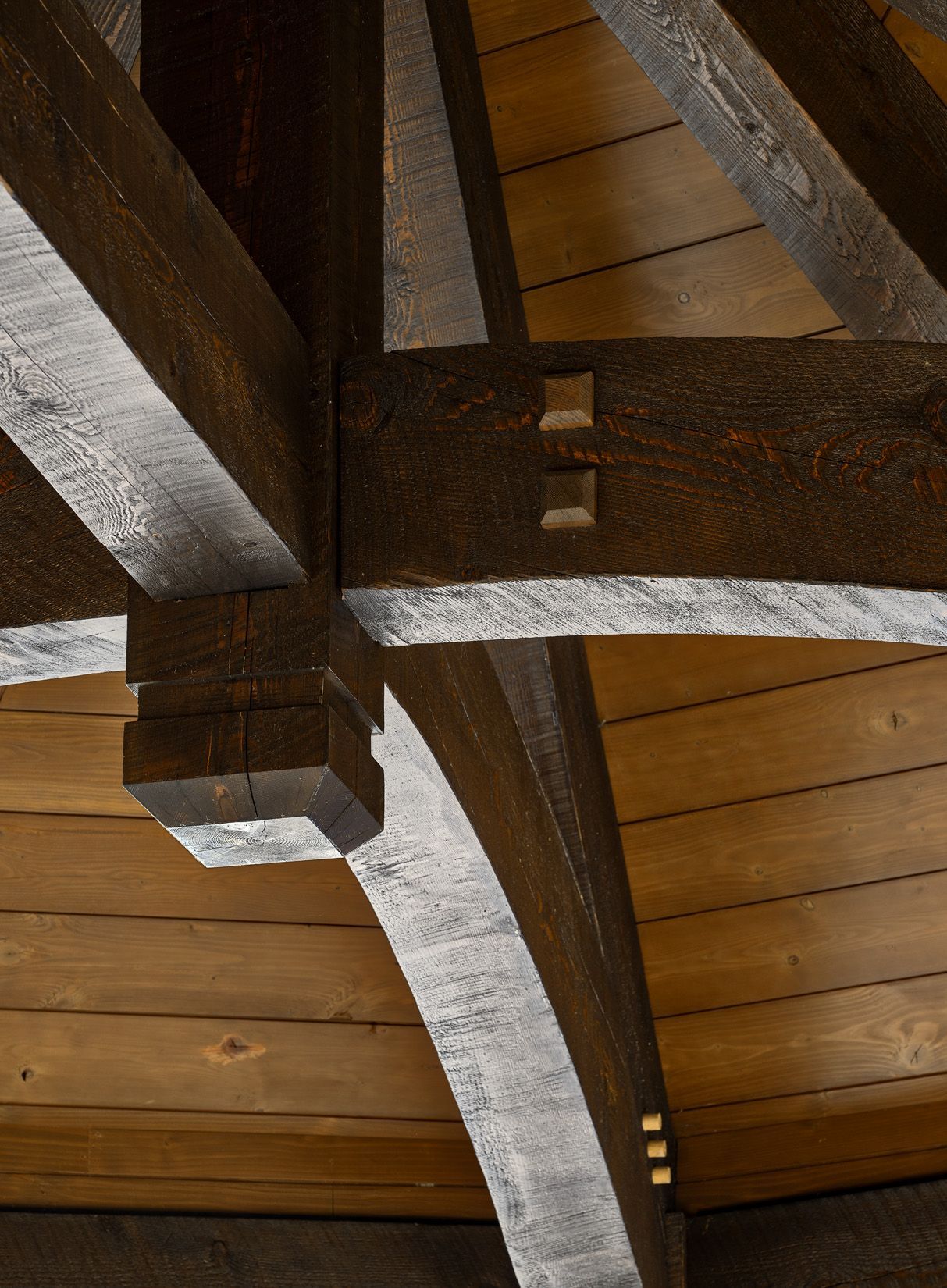 Close-up of heavy dark wood timber ceiling beams joined together with wooden pegs, set against light wood planking.