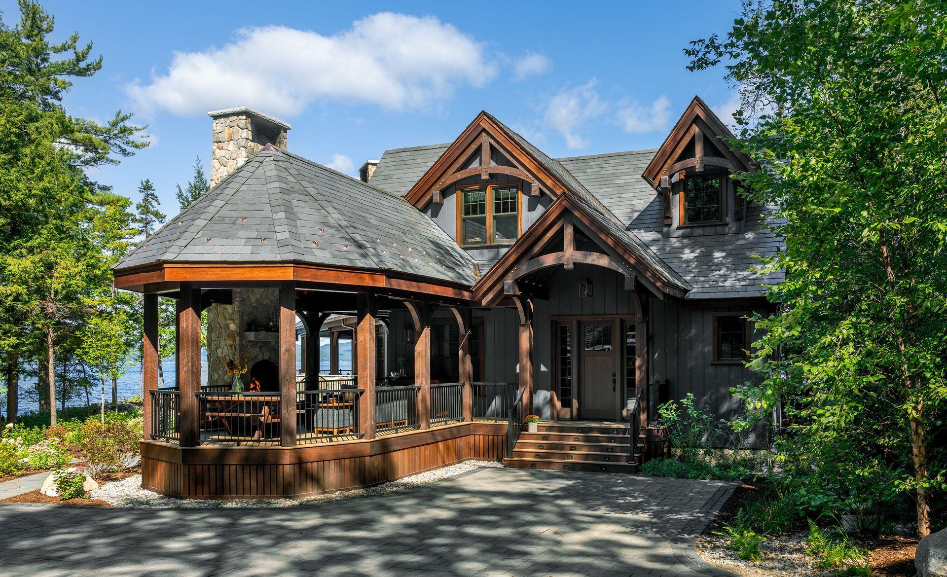 A rustic house with a dark wood exterior, stone chimney, and a sprawling, covered porch nestled in a lush, green forest.