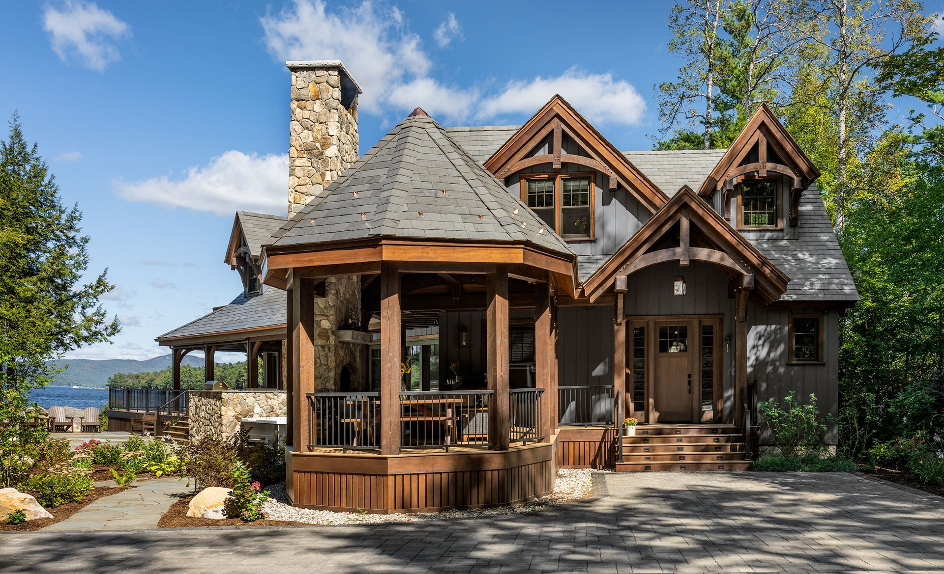 A rustic, multi-story home with a stone chimney, a hexagonal porch, and wood-framed gables, set against a wooded landscape.