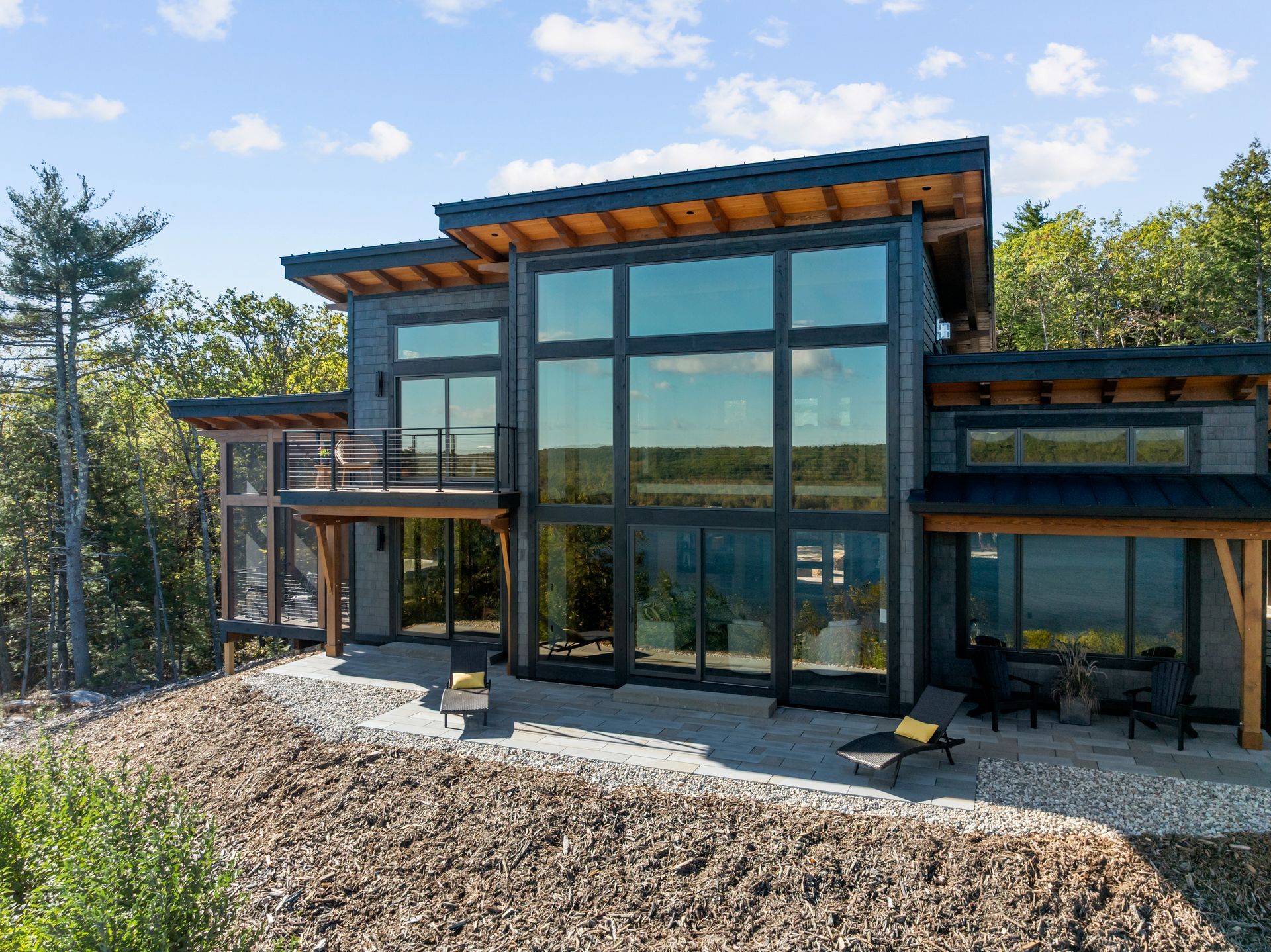 A modern, gray, multi-story house with large floor-to-ceiling windows, a balcony, and a stone patio in a wooded setting.