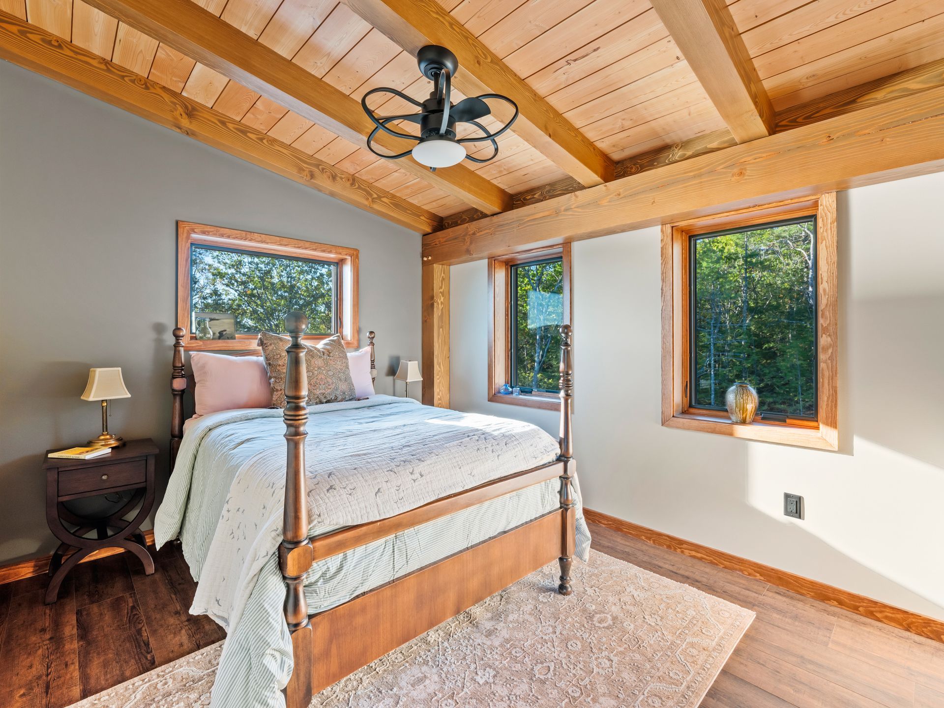 A cozy bedroom with a wooden four-poster bed, wood-paneled ceiling, two windows, and a dark side table with a lamp.