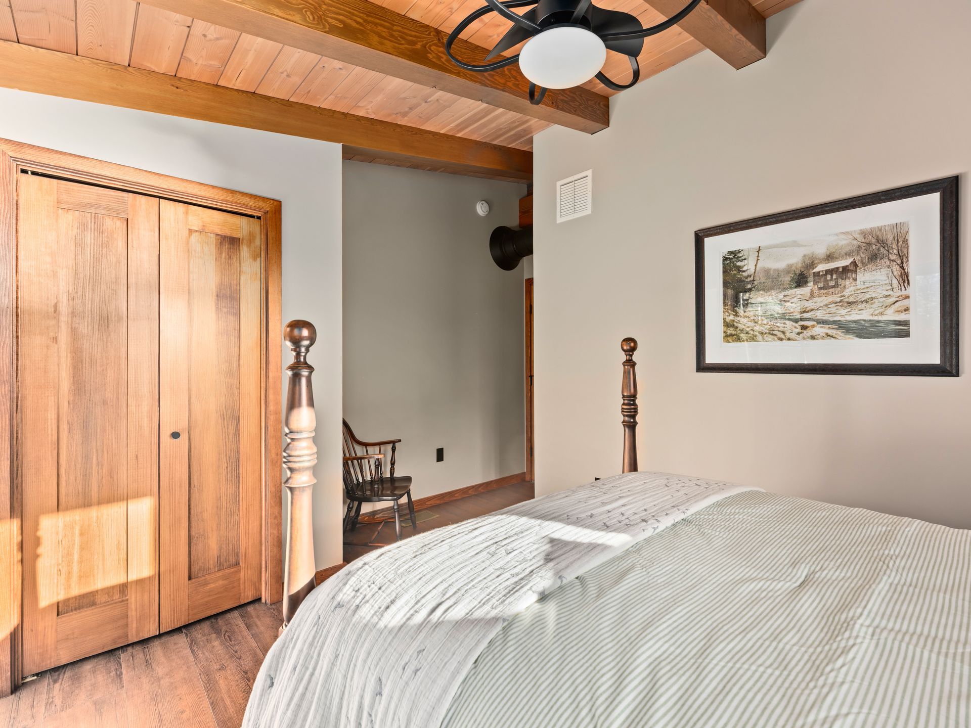 A bedroom with a bed featuring a striped comforter, wooden closet doors, exposed ceiling beams, and wall art.