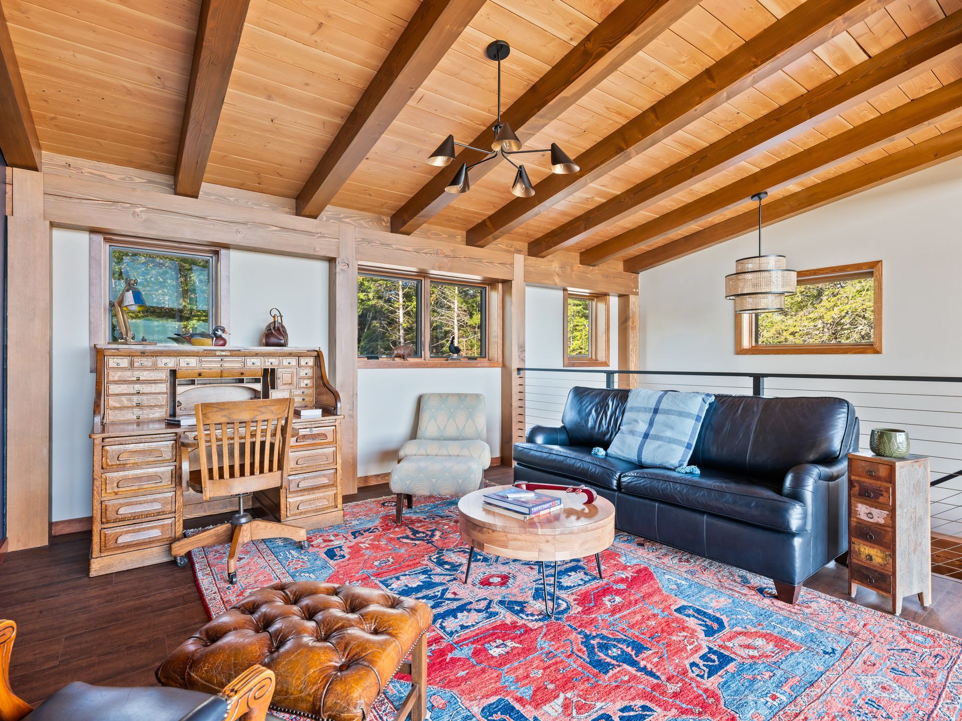 A living room with exposed wood beams, a desk, leather sofa, patterned area rug, and wood-paneled ceiling.