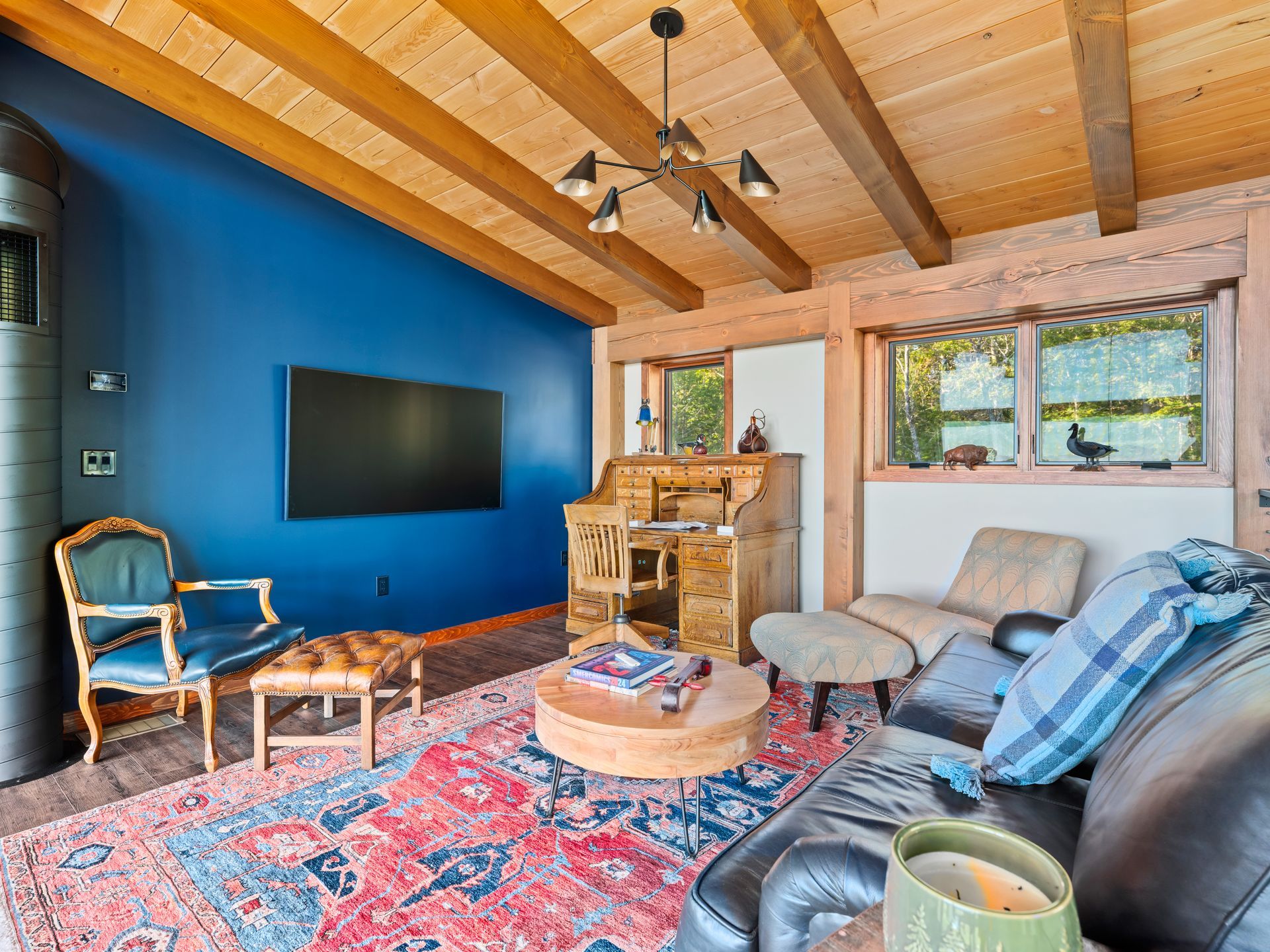 A cozy living room with a bright blue accent wall, a TV, a patterned rug, wooden beams, and diverse seating furniture.