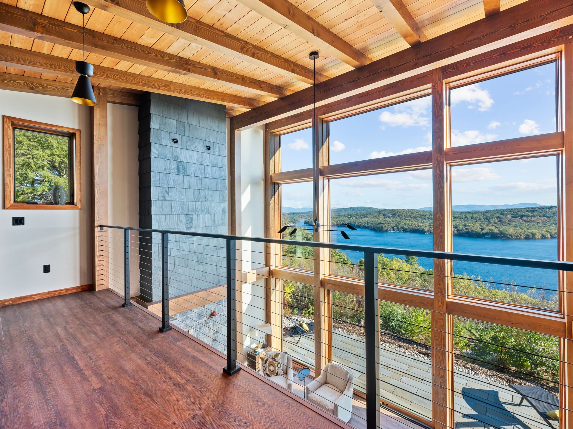 A loft area with wood flooring and ceiling beams overlooks a large window with a scenic lake and forest view.