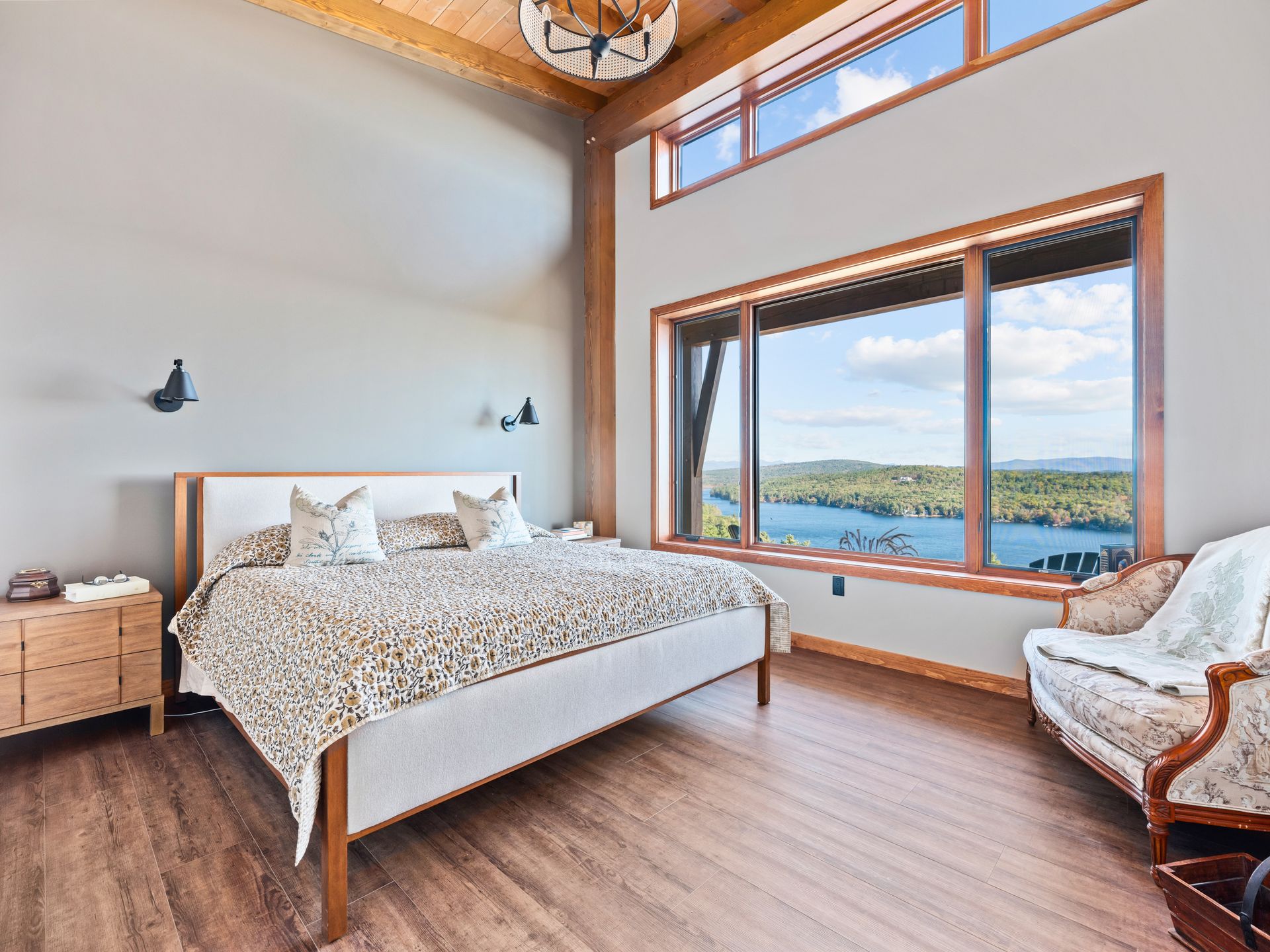 A bedroom with a king-sized bed, wood floors, and large windows offering a scenic view of a lake and forest.
