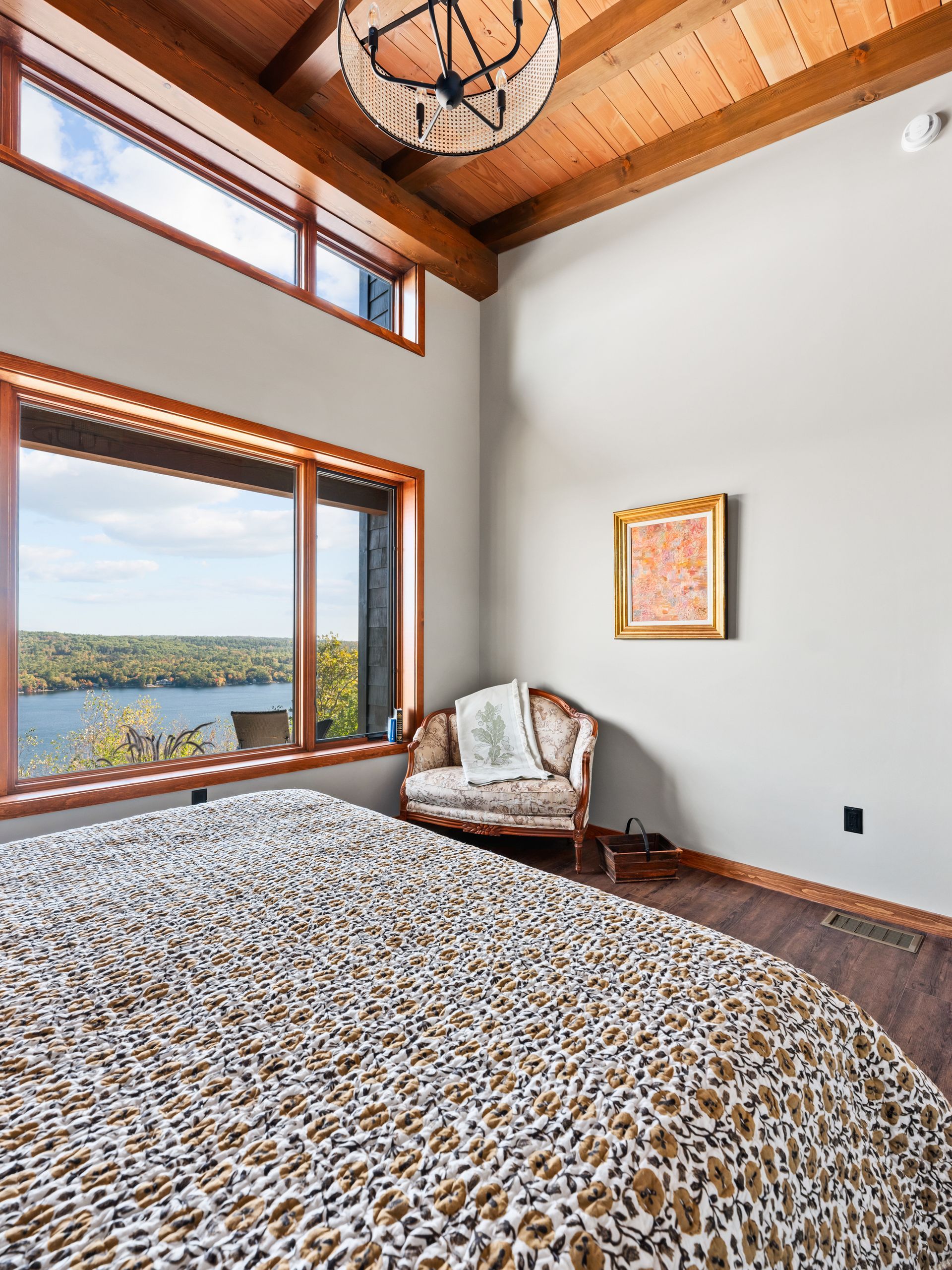 A bedroom with a high wood-beam ceiling, a view of a lake through large windows, and an upholstered armchair.