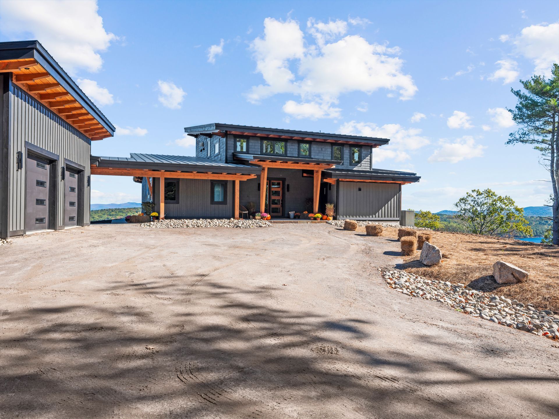 A modern, gray, multi-level house with wood beams and a large gravel driveway under a bright blue sky.