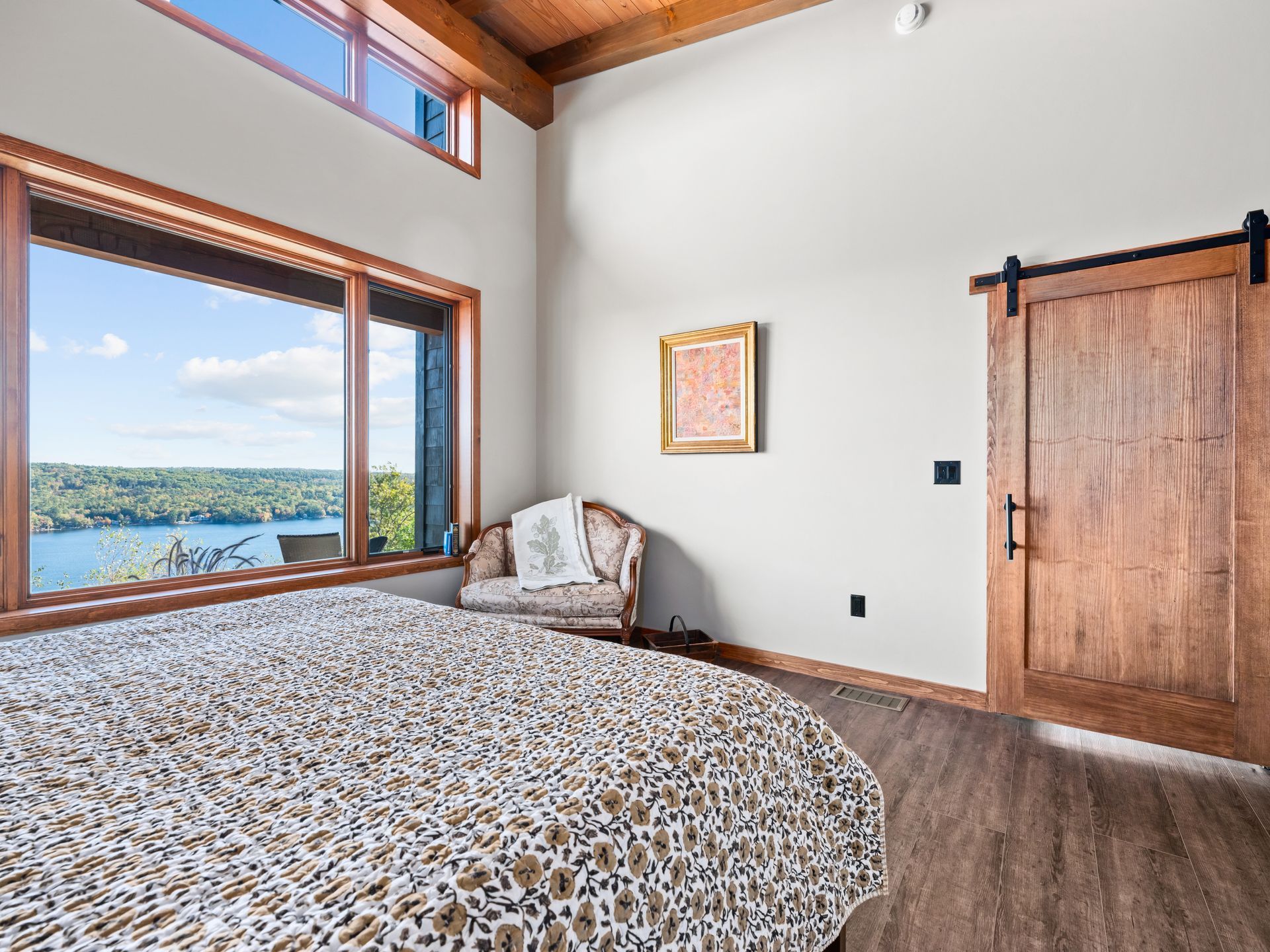 Bedroom with a wooden barn door, a large window overlooking a lake, and a patterned bedspread under a sloped wood ceiling.