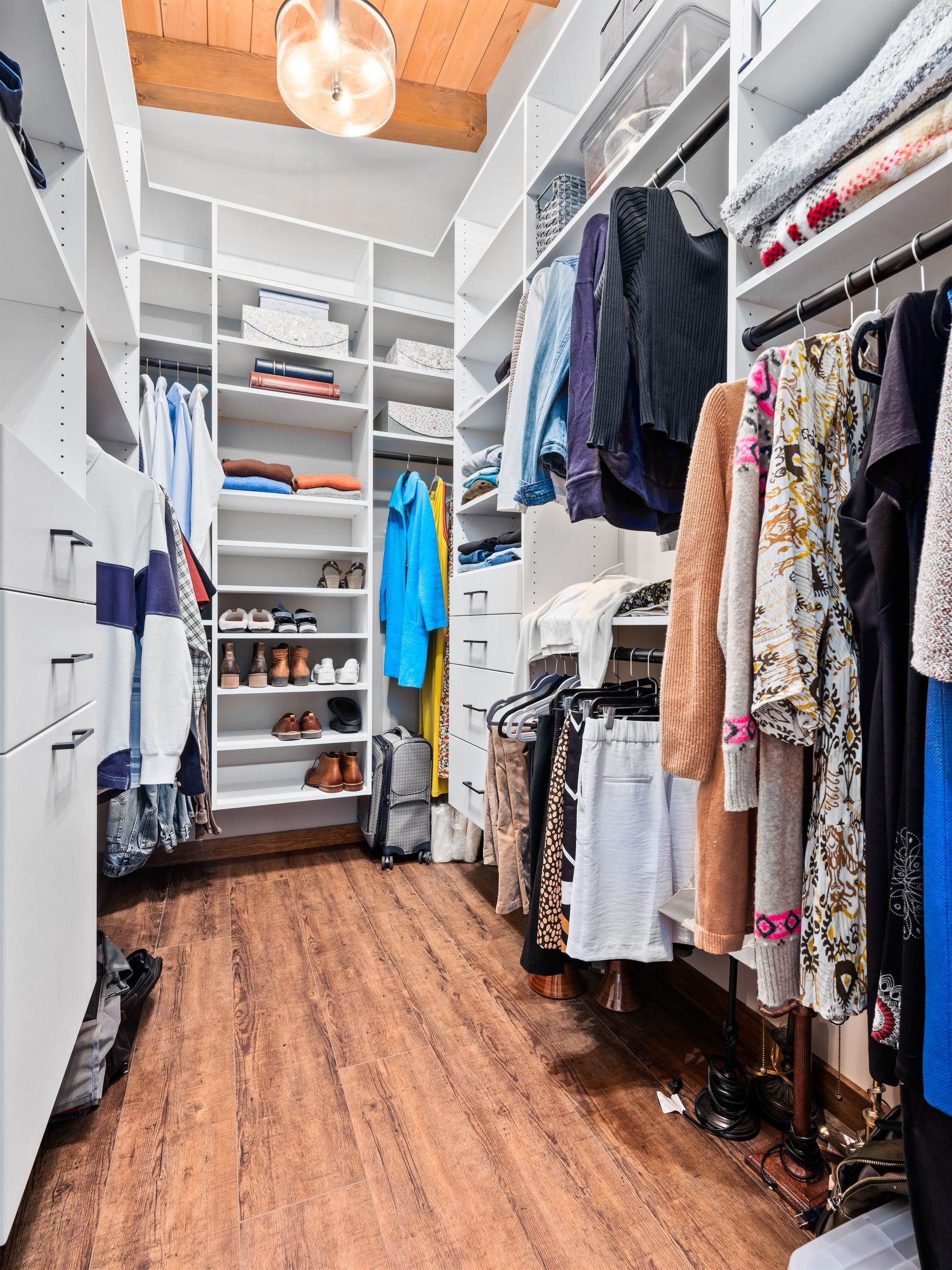A walk-in closet with white shelving, hanging clothes, shoes, and wooden flooring under a bright light fixture.
