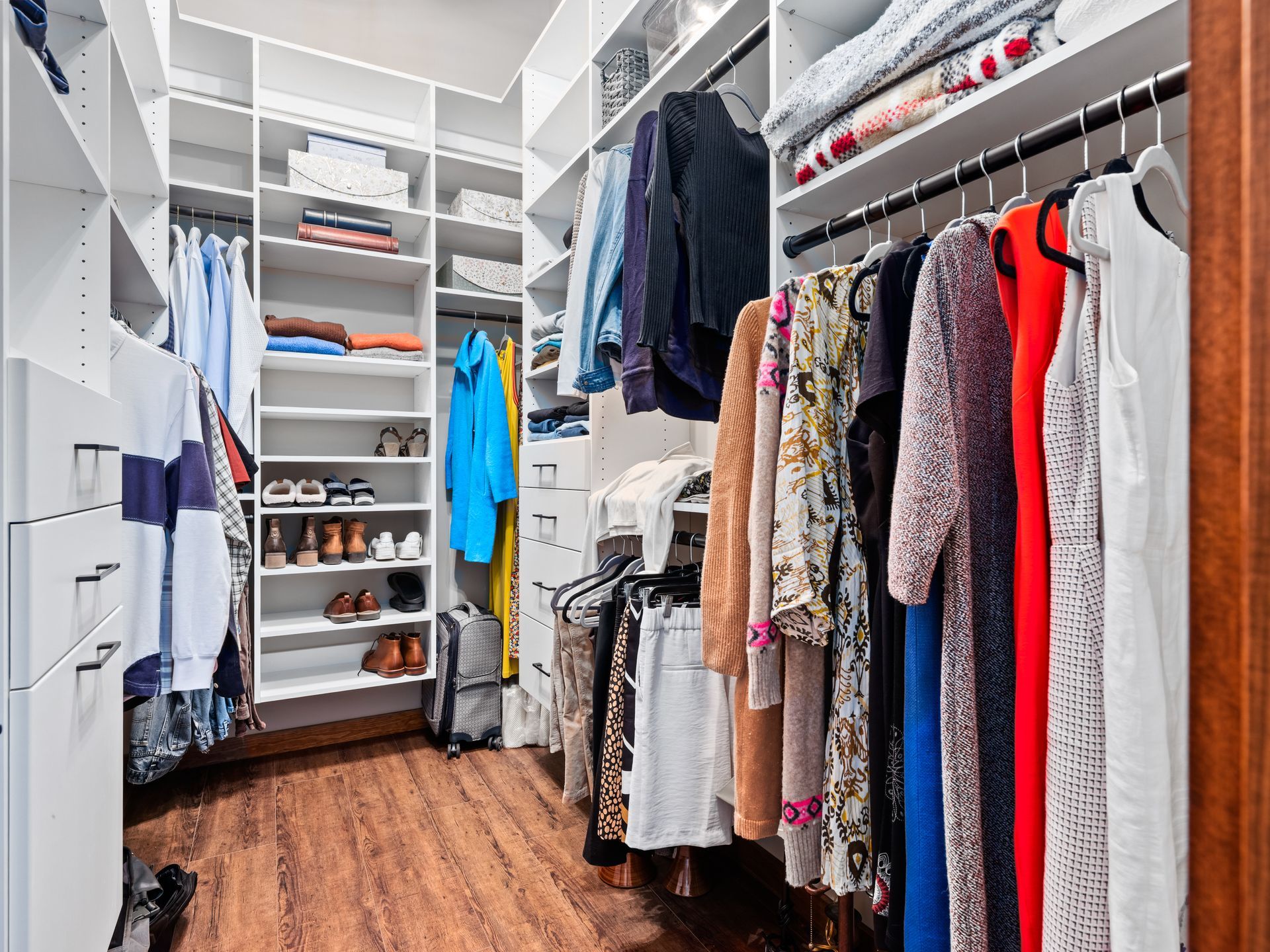A well-organized walk-in closet with white shelving, hanging clothes, and wood-look flooring.