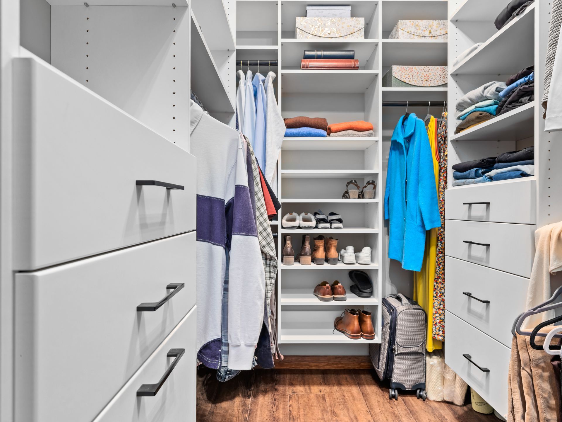 A bright, modern walk-in closet featuring white shelves, hanging clothes, organized shoes, and drawers with dark hardware.