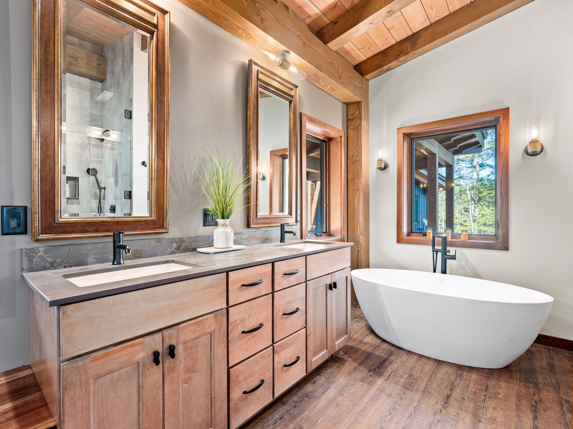 A rustic-style bathroom with a double wood vanity, matching framed mirrors, wood-look flooring, and a white soaking tub.