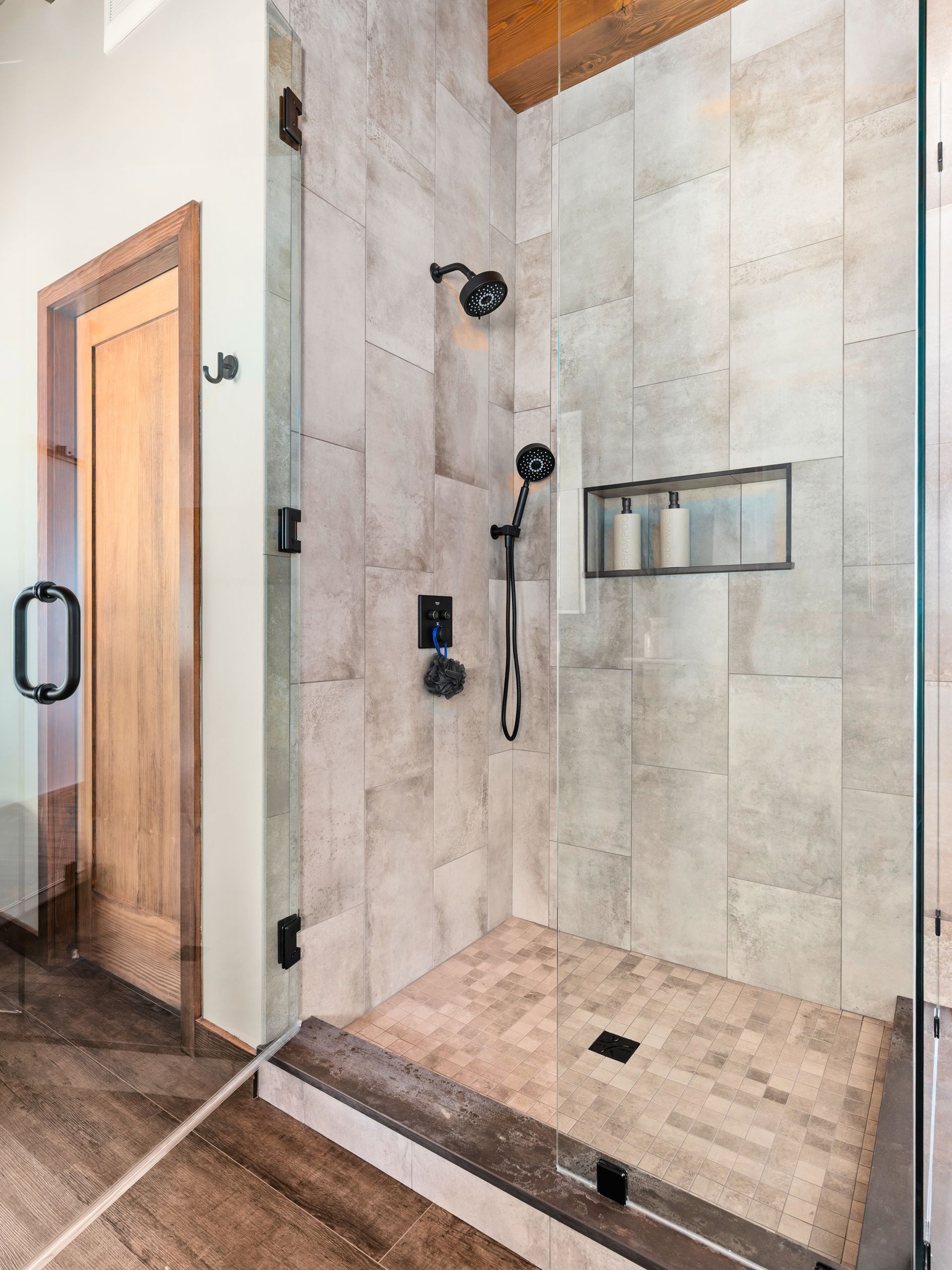 A modern walk-in shower with light stone-look wall tiles, a mosaic tiled floor, black fixtures, and a glass door.