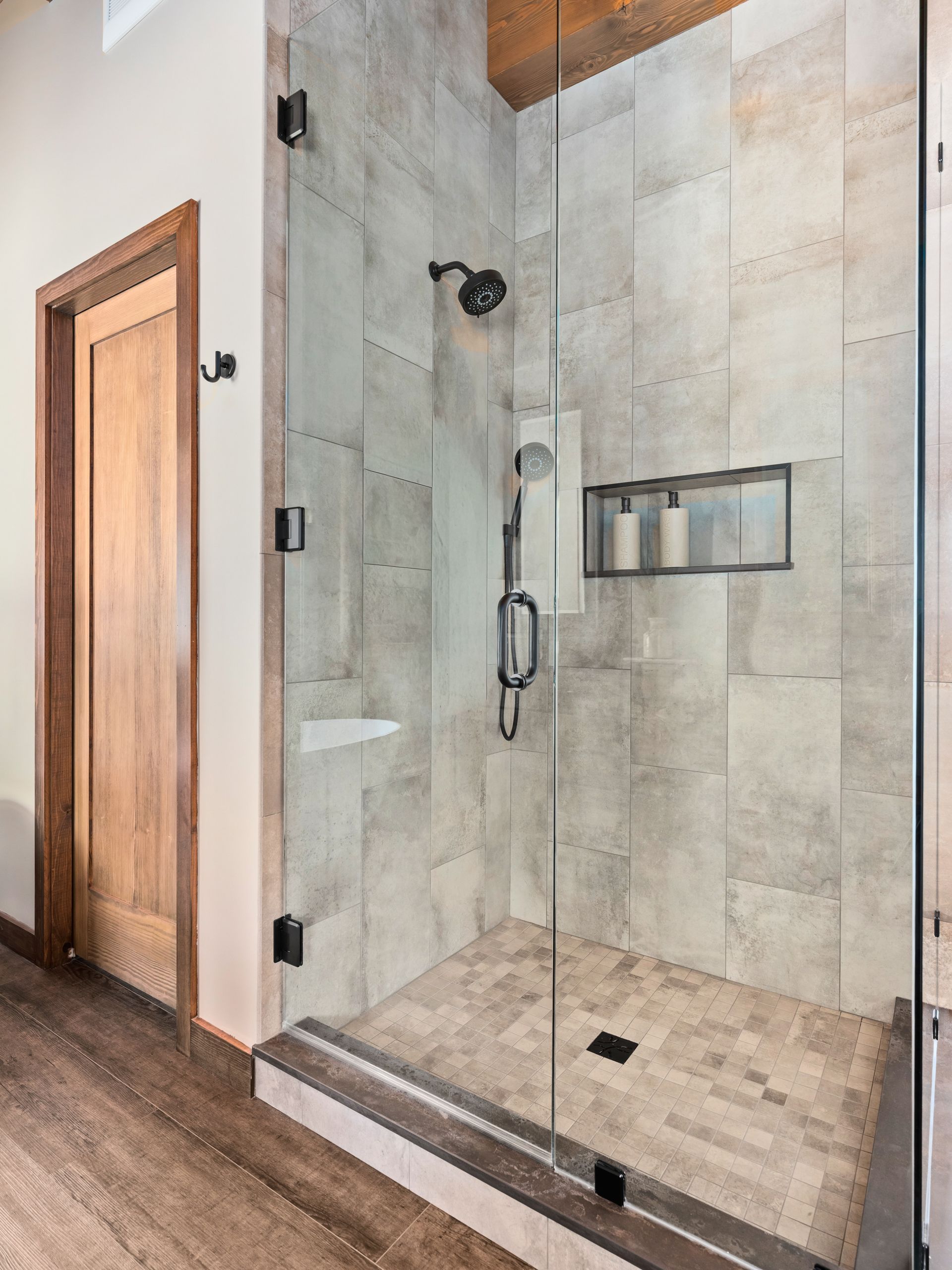 A walk-in shower with light stone-look tiles, a glass door, a black rainfall showerhead, and a recessed wall niche.