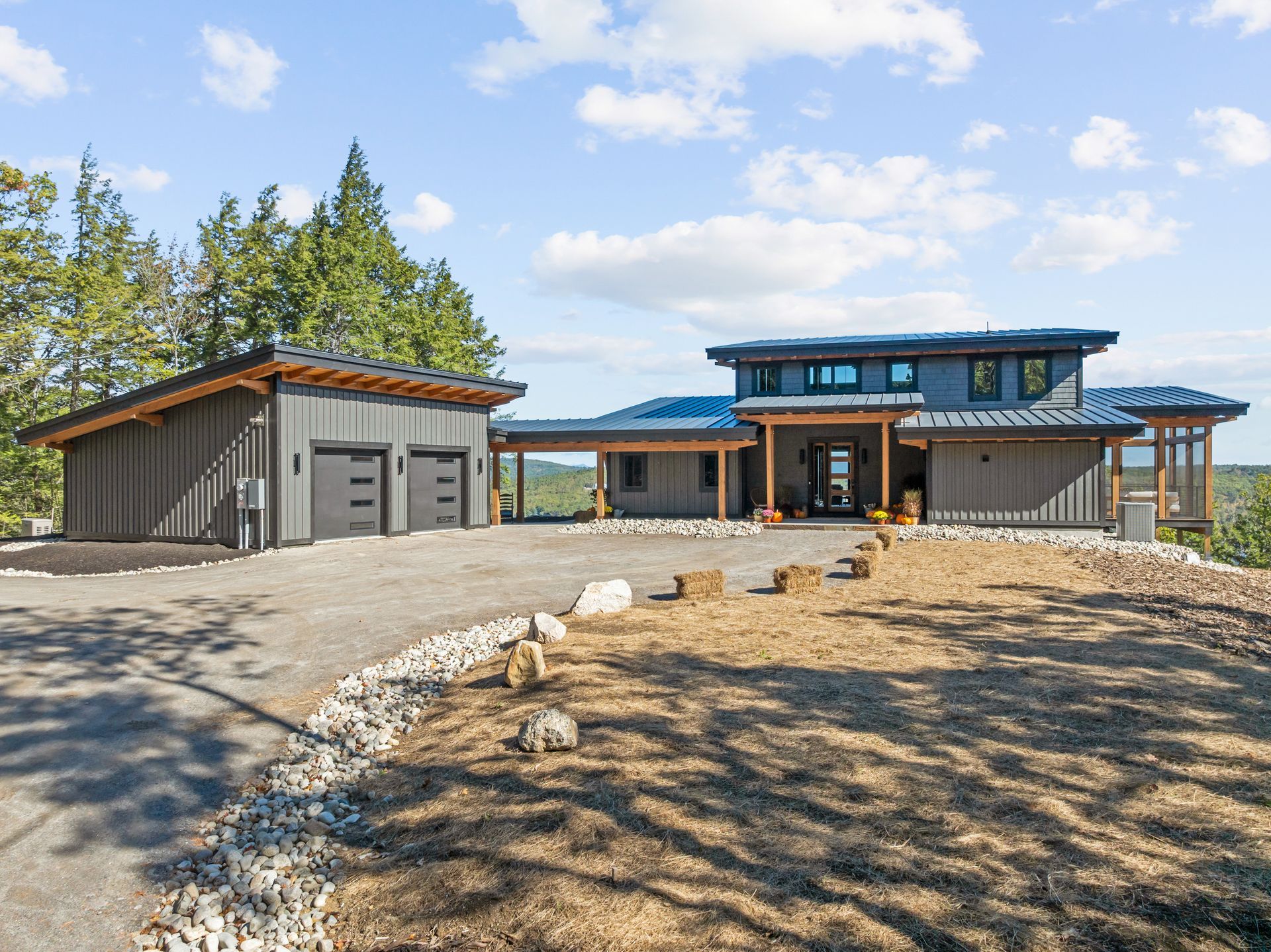 A modern dark-gray house with a flat roof, a two-car garage, and a large gravel driveway in a wooded, sunny setting.