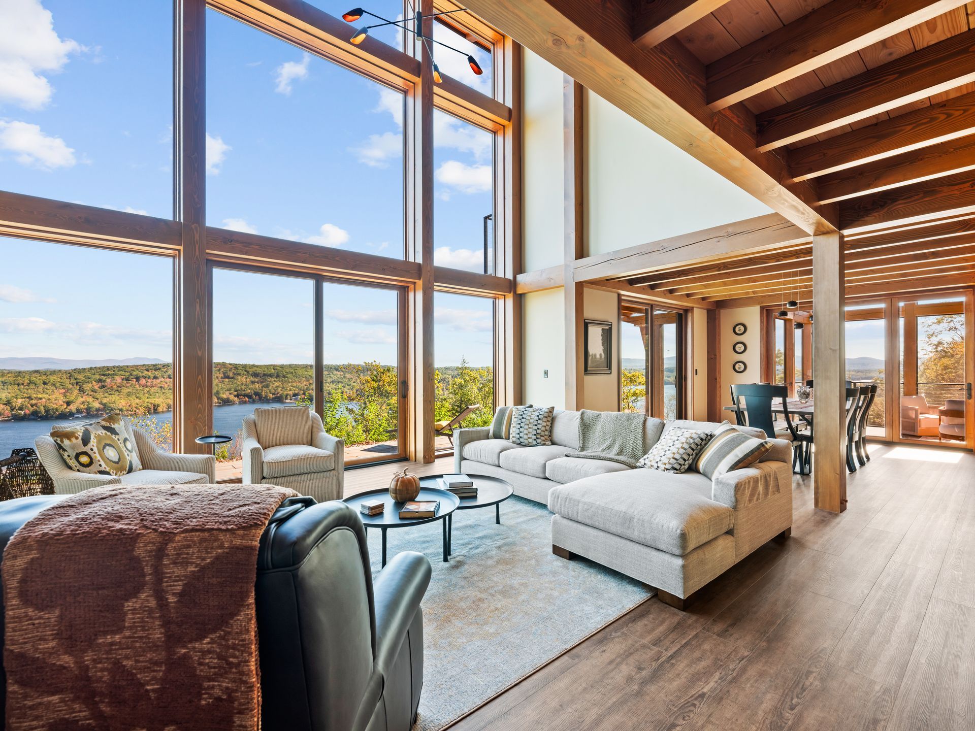 Spacious, modern living room with floor-to-ceiling windows overlooking a lake and forest, wooden beams, and neutral seating.