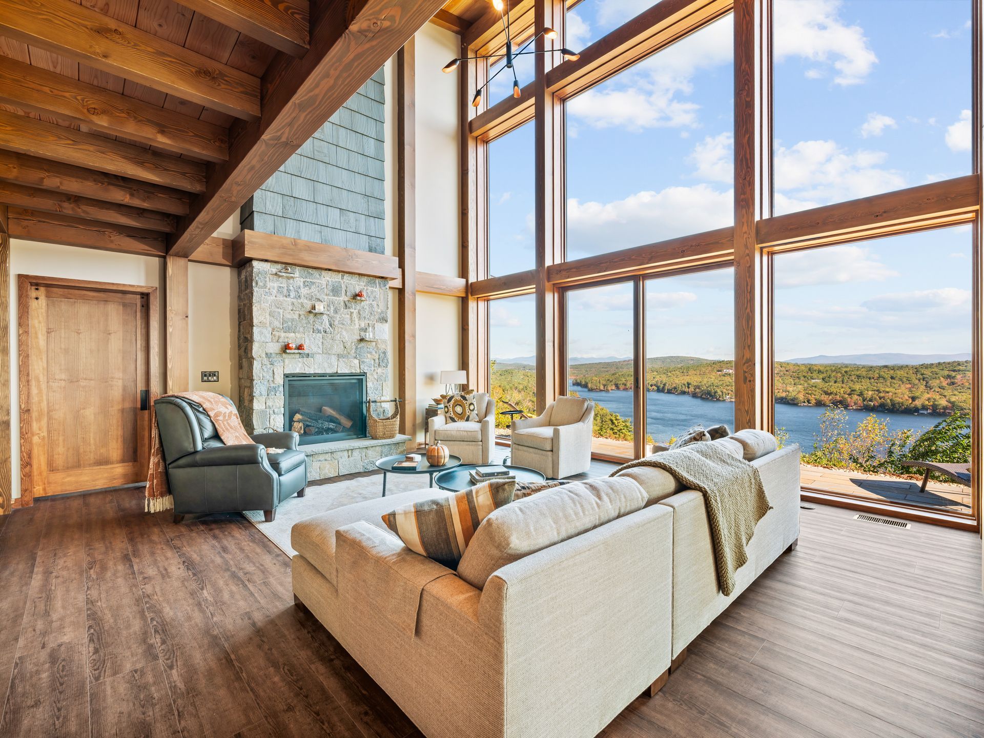 A bright, spacious living room with large windows overlooking a lake, stone fireplace, and modern neutral-toned furniture.