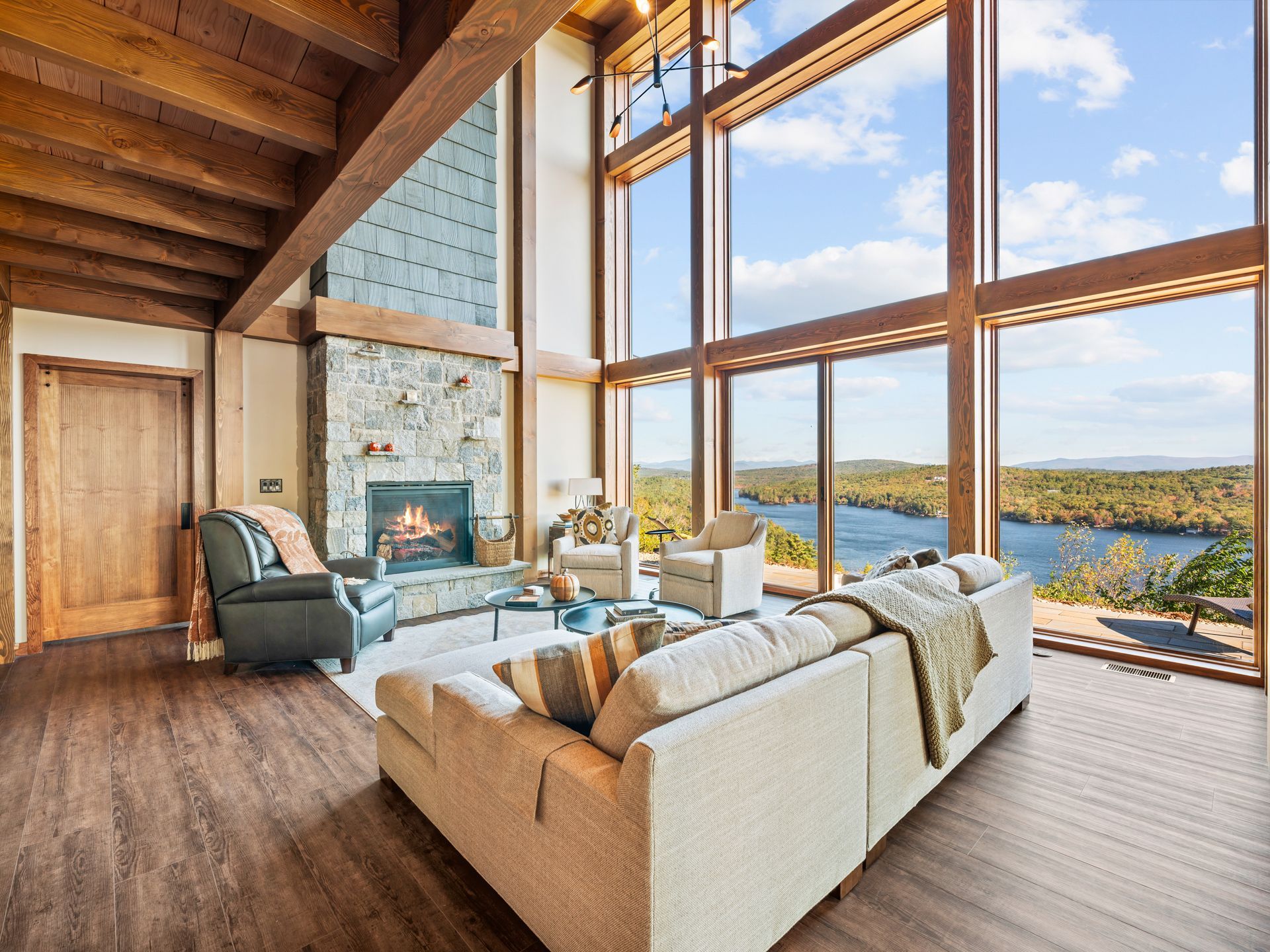 A modern, high-ceilinged living room with a large stone fireplace and expansive windows overlooking a river and woods.