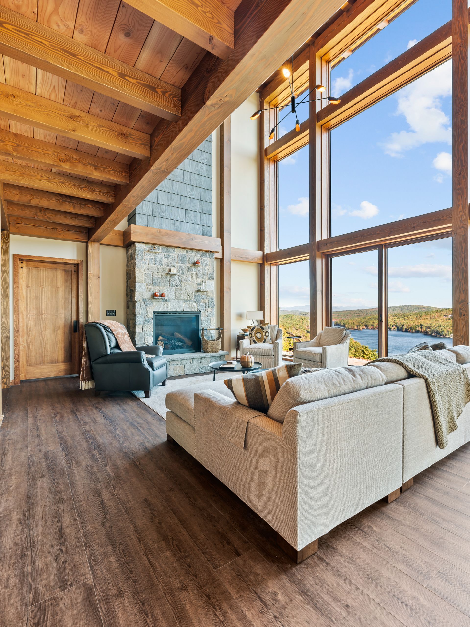 A cozy living room with high beamed ceilings, a stone fireplace, and large windows overlooking a lake.