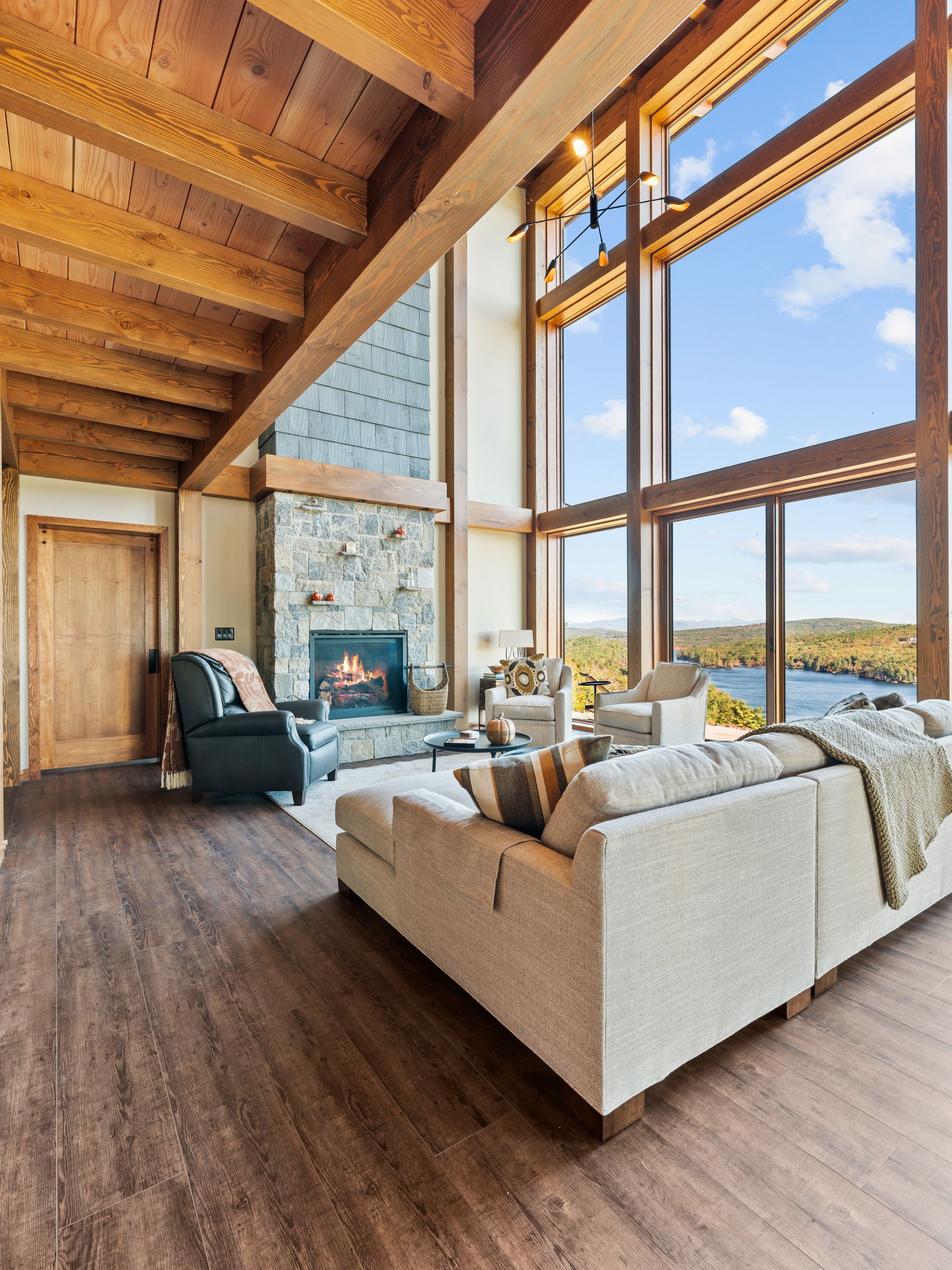 A cozy living room with exposed wood beams, a stone fireplace, and large windows overlooking a lake and forest.