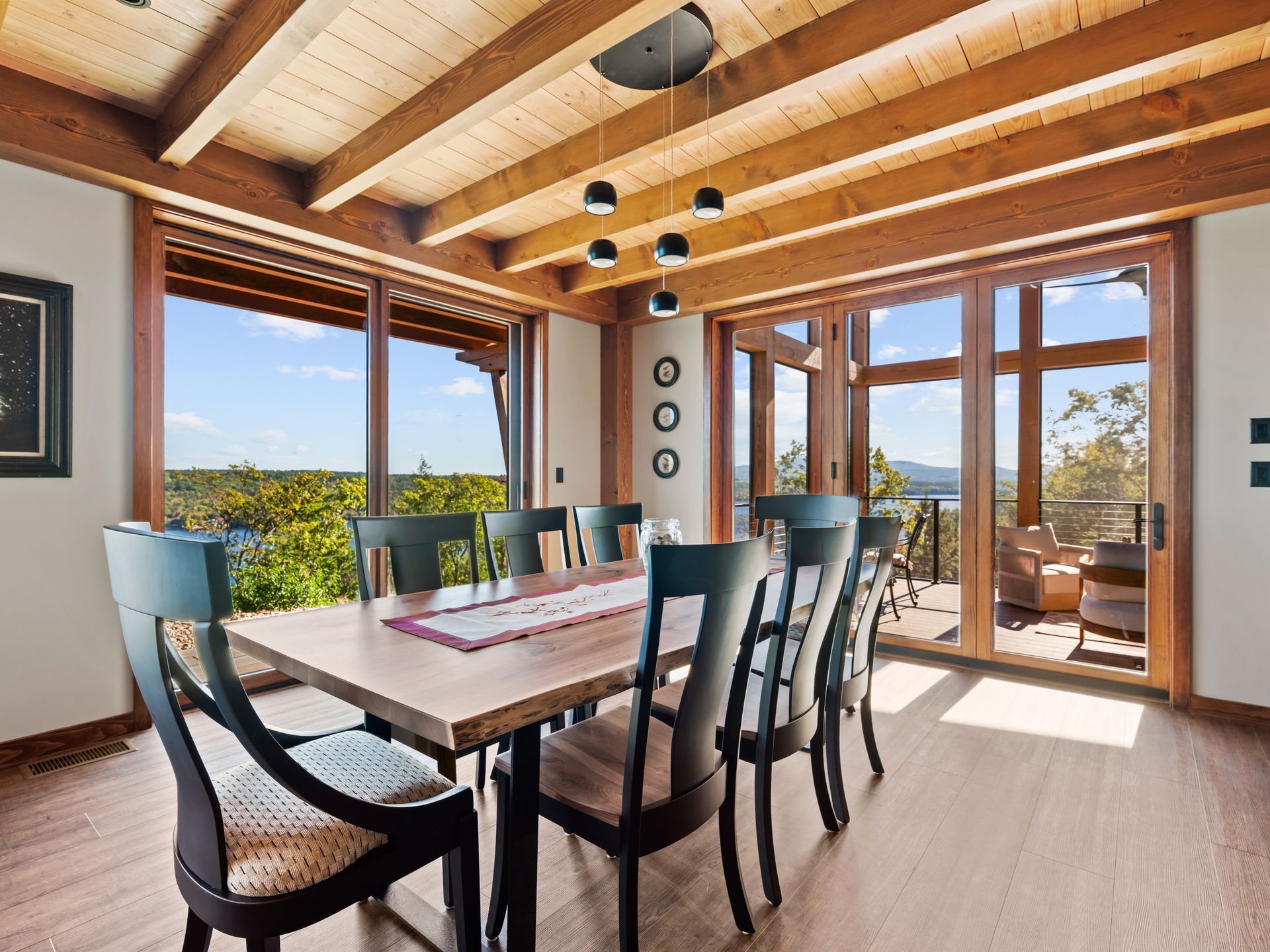 A modern dining room with a long wooden table, six dark chairs, wood-beam ceiling, and large windows overlooking trees.