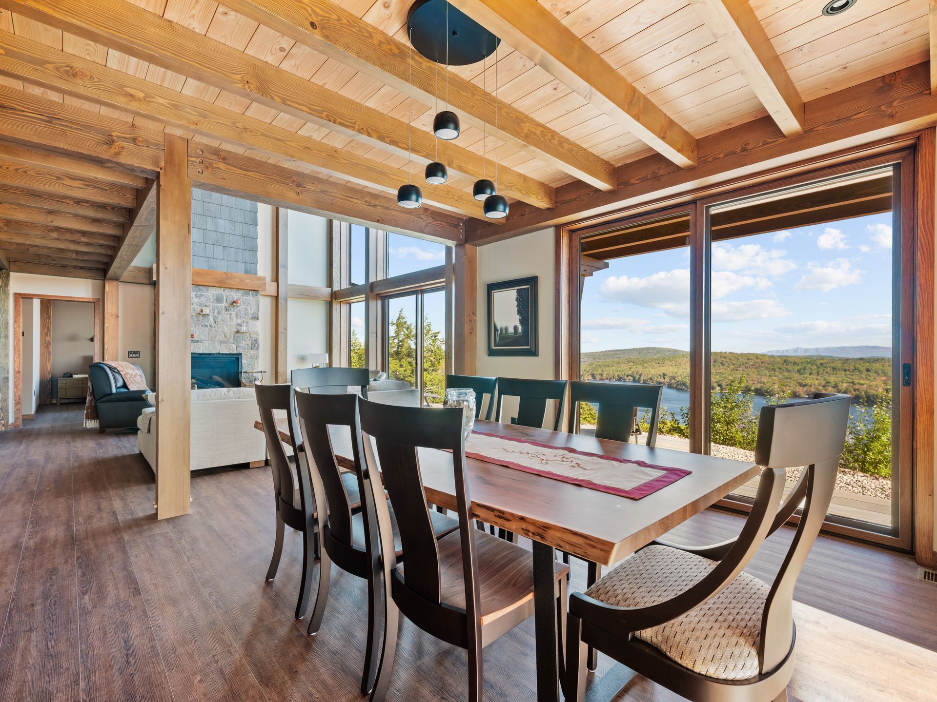 A wooden dining table with black chairs sits in a bright, modern room overlooking a scenic forest and water view.