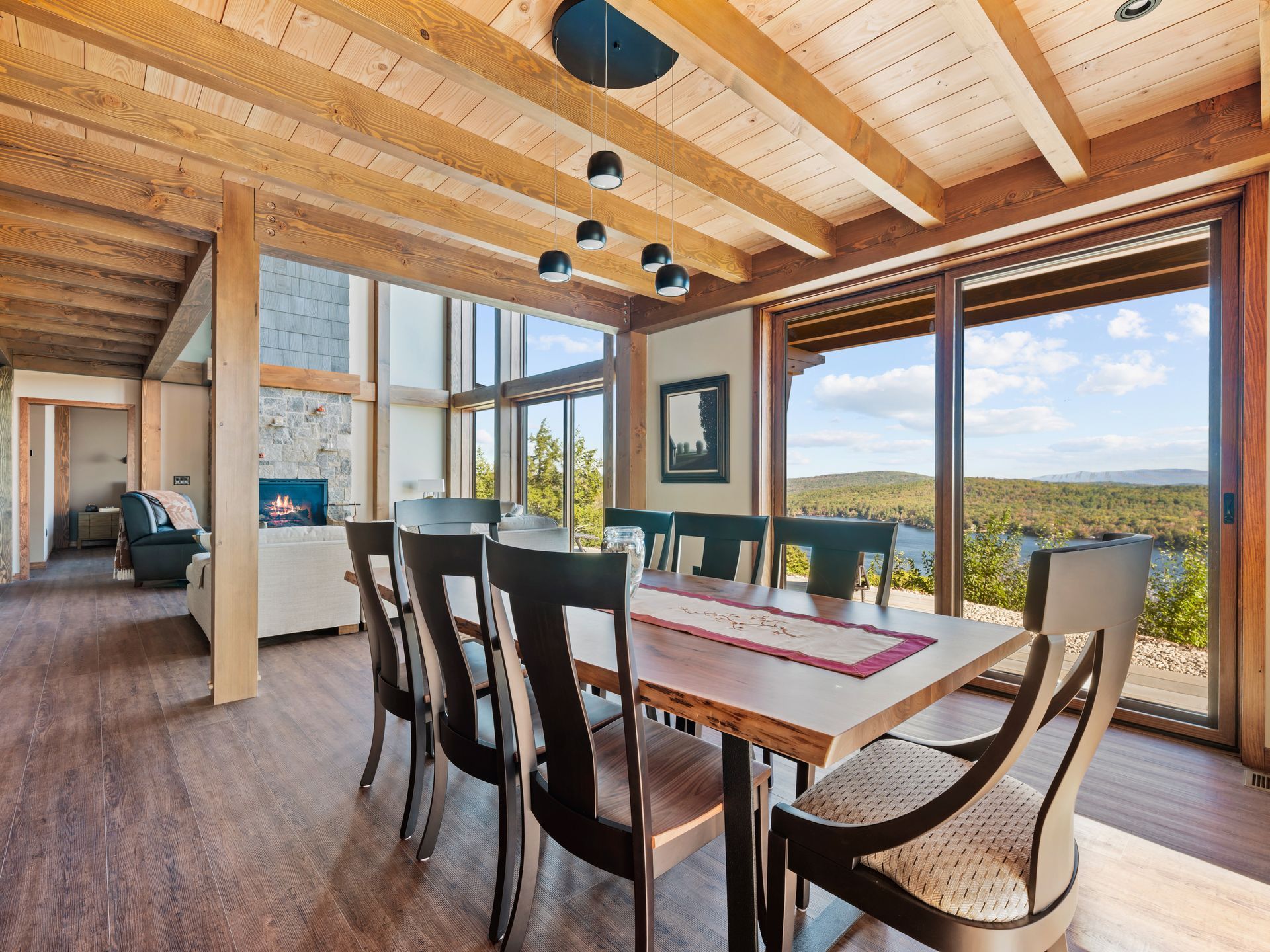 A wooden dining set sits under ceiling beams in a room with floor-to-ceiling windows overlooking a forest and lake.