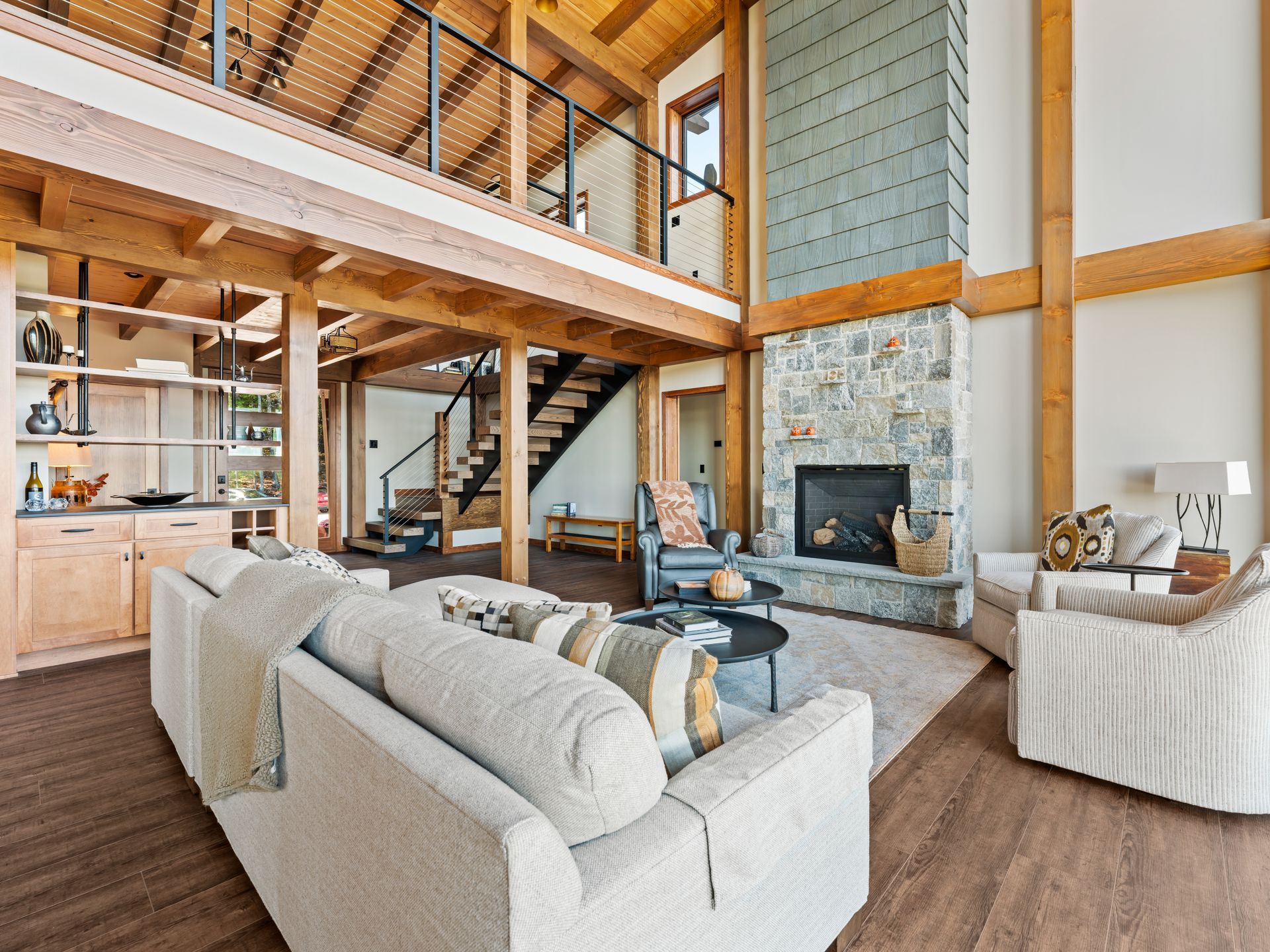 A spacious, high-ceilinged living room with wood beams, a stone fireplace, neutral sofas, and a wooden staircase.
