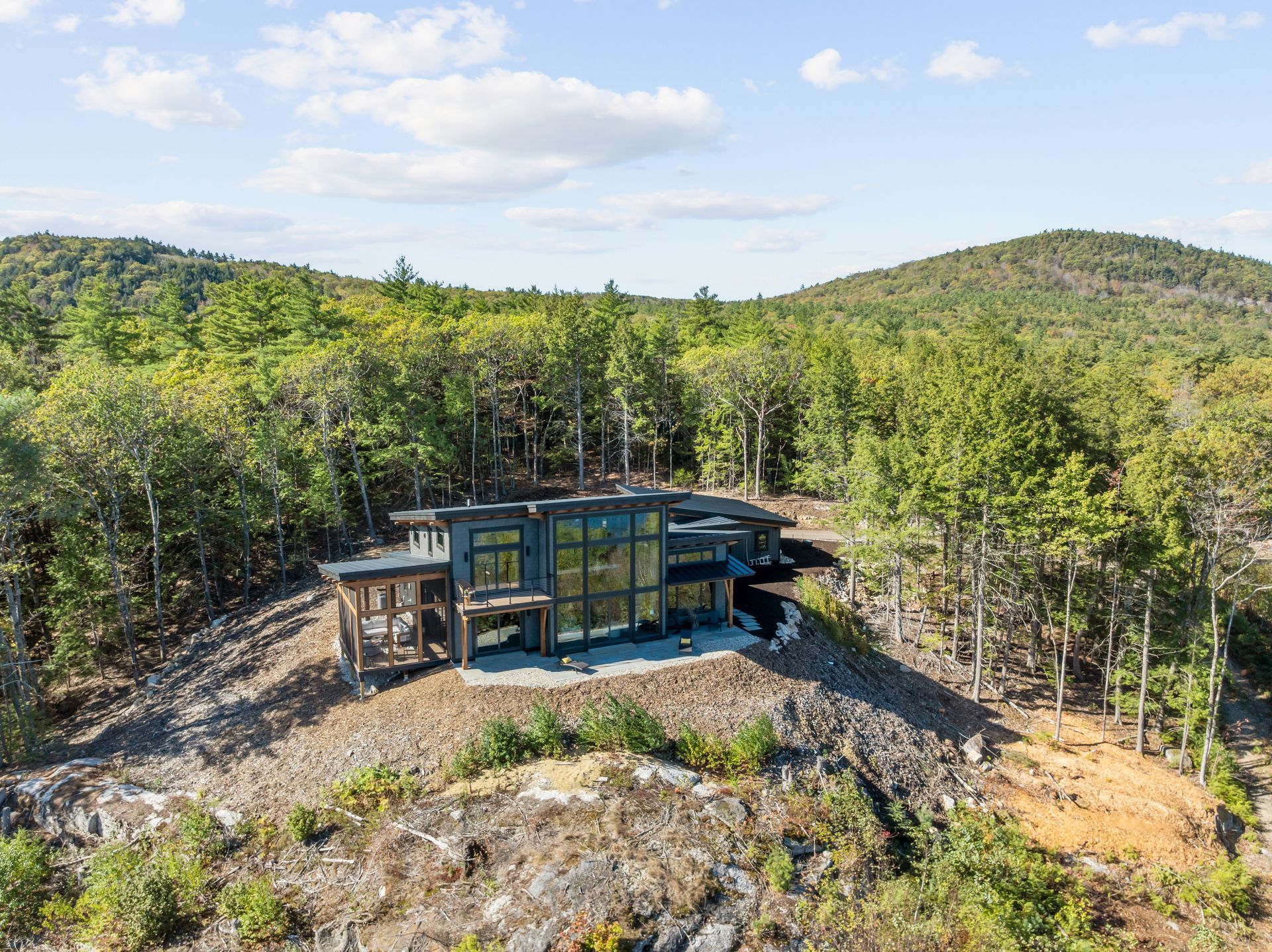 An aerial view of a contemporary, glass-walled modern house nestled atop a rocky, tree-covered hill under a blue sky.