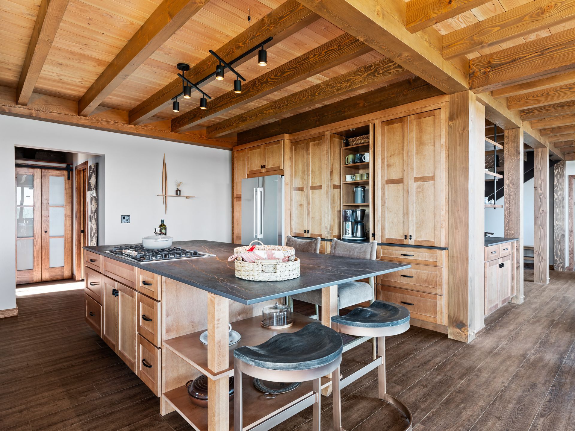 A rustic kitchen featuring a large slate-topped island with wooden stools, light wood cabinetry, and exposed ceiling beams.