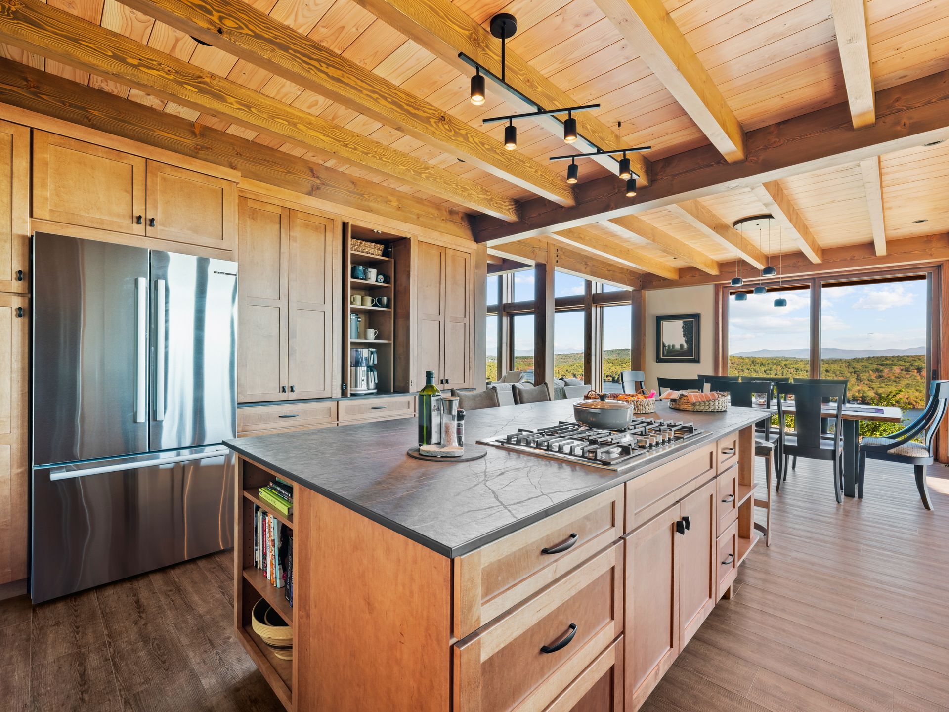A bright, modern kitchen with light wood cabinetry, a large central island with a stove, and large windows with a view.