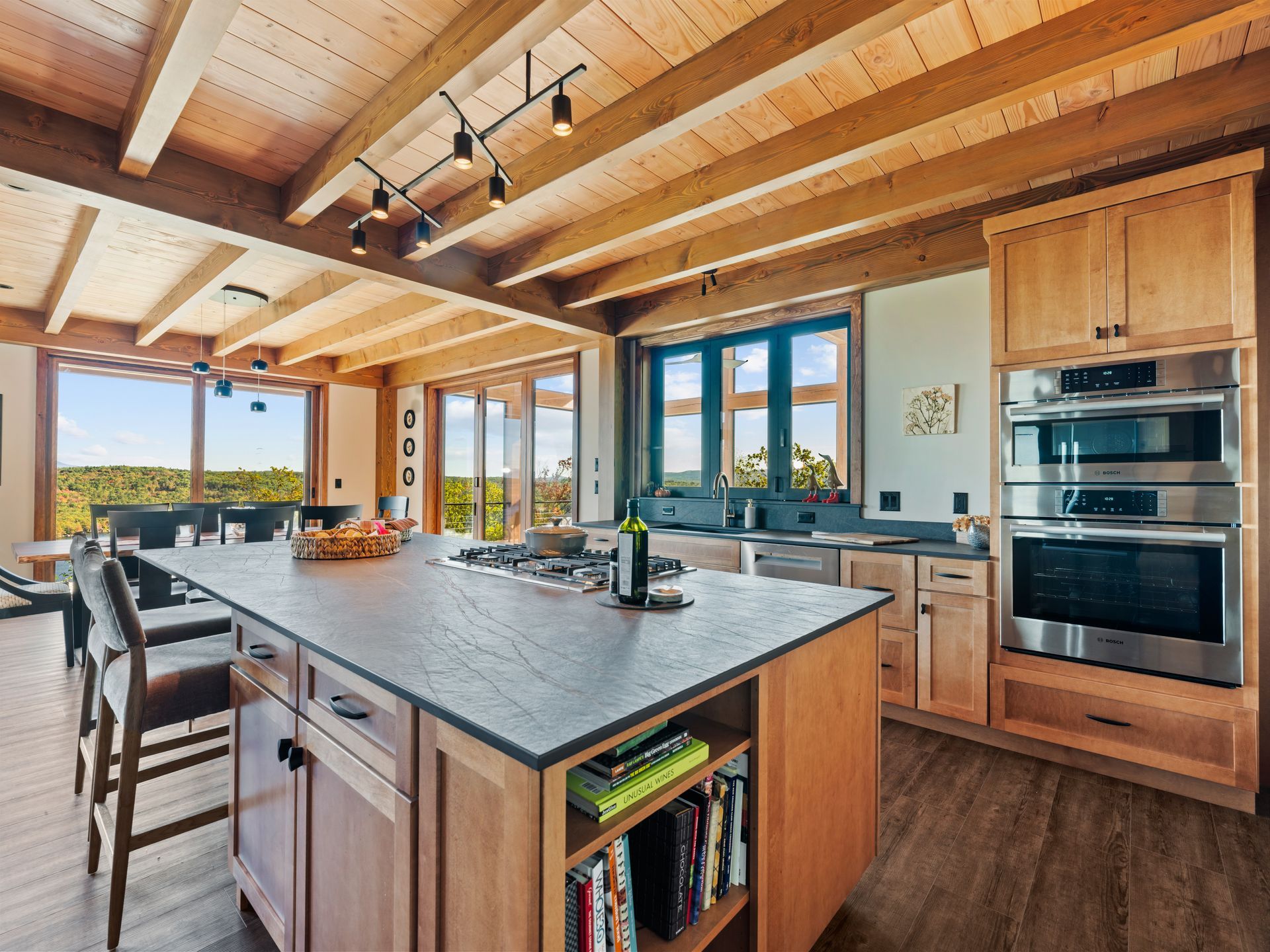 A modern kitchen with a large island, wooden cabinetry, beamed ceilings, and a wall-mounted double oven.