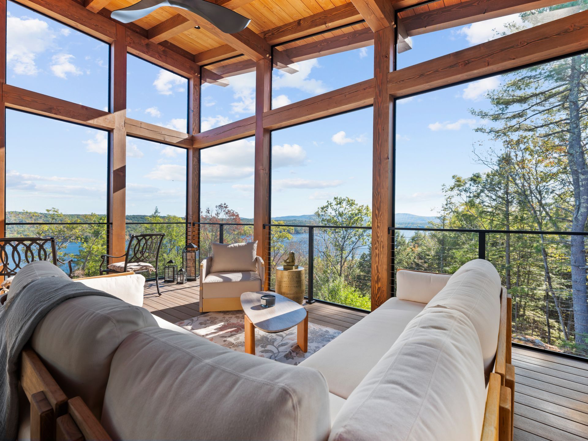 A cozy, screened-in porch with light-colored sofas, a wooden frame, and expansive windows overlooking a scenic, wooded view.