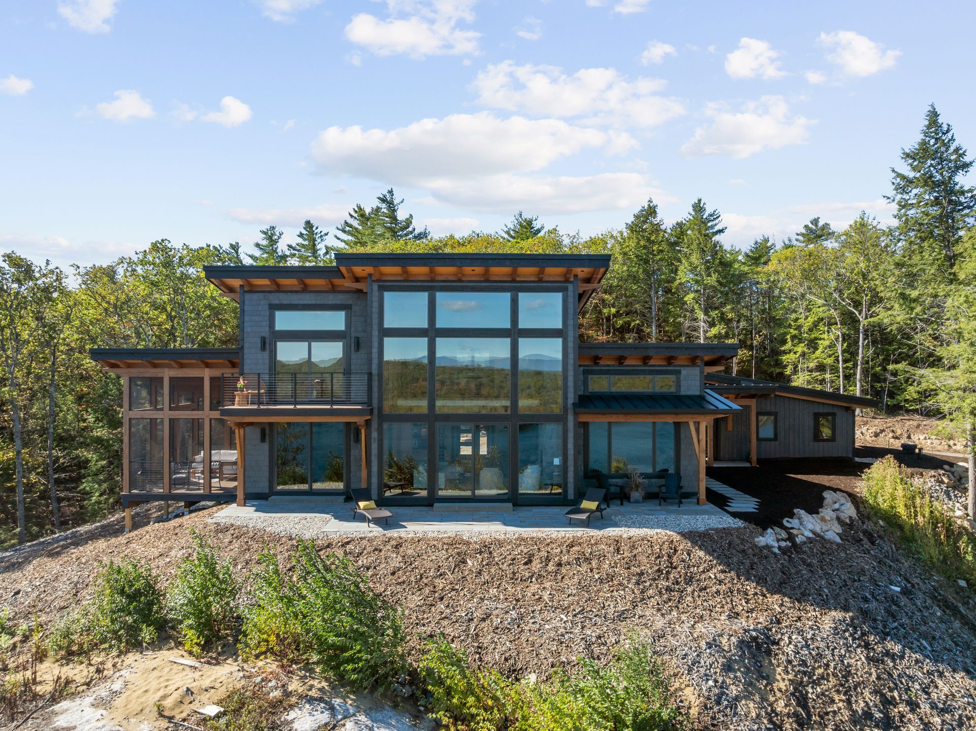 A modern two-story home with expansive glass windows and a stone patio, set on a rocky hillside surrounded by forest.