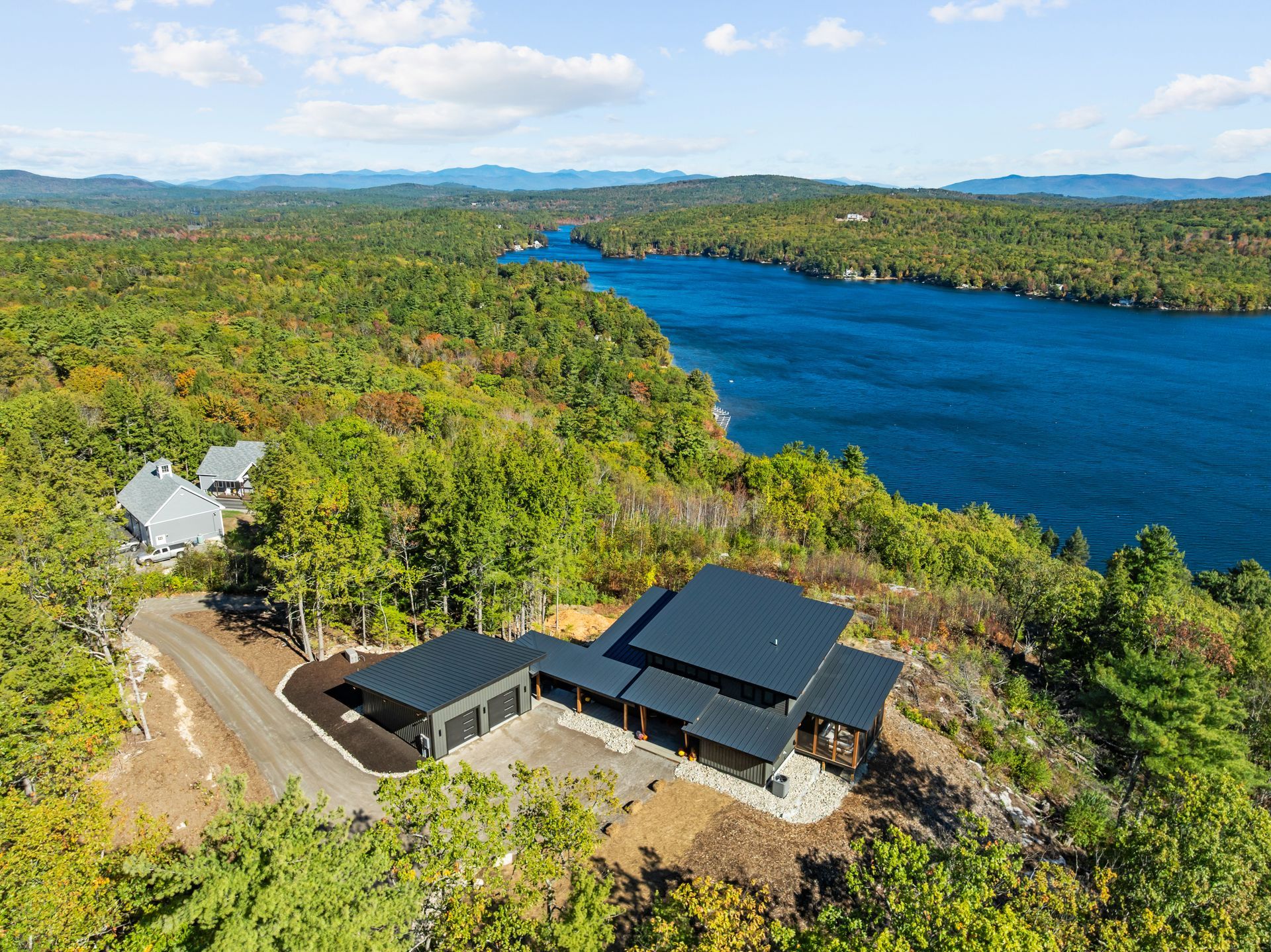 An aerial view of a modern dark-roofed house nestled in a dense green forest next to a large blue lake.
