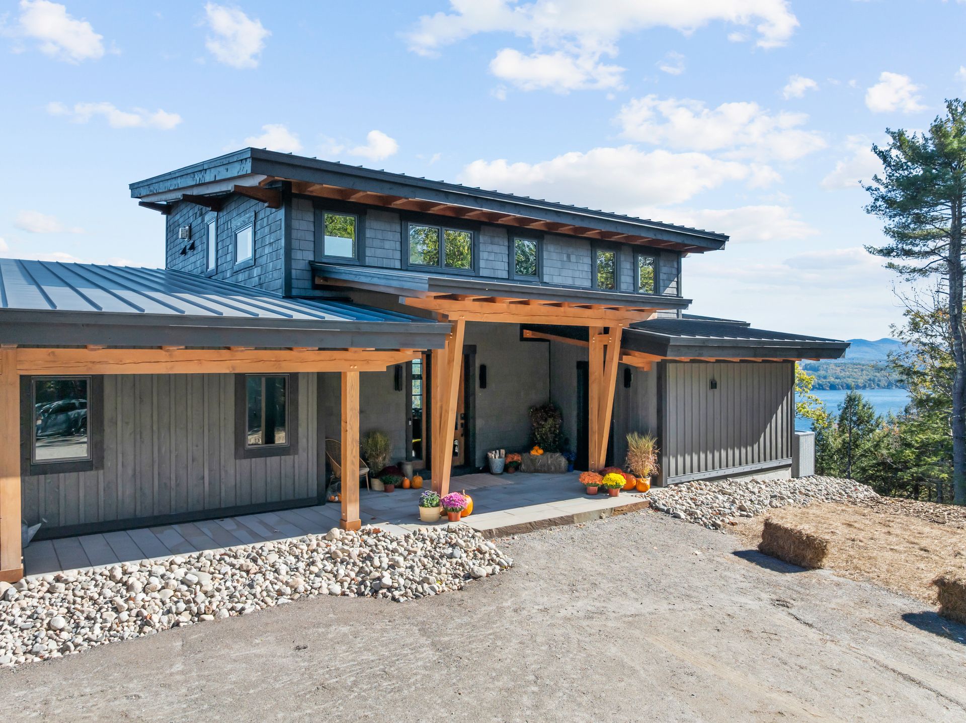 A modern home with grey vertical siding and metal roofing, featuring a timber-framed entry and a gravel driveway.
