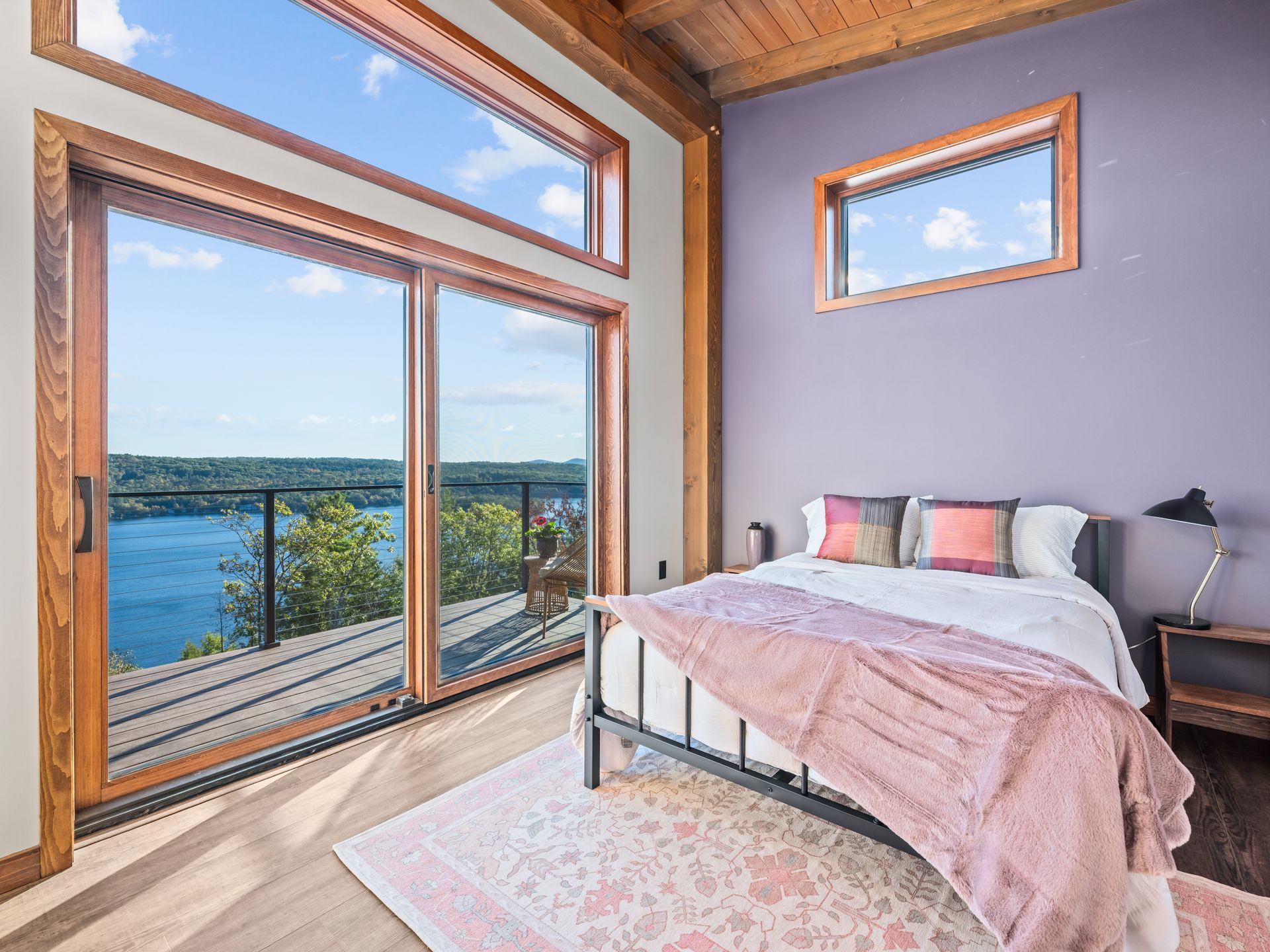 A bedroom with a queen bed, pink bedding, and floor-to-ceiling windows overlooking a blue lake and surrounding trees.