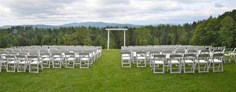Rows of white chairs face a wedding arch on a grassy field, with a forest and mountain backdrop.