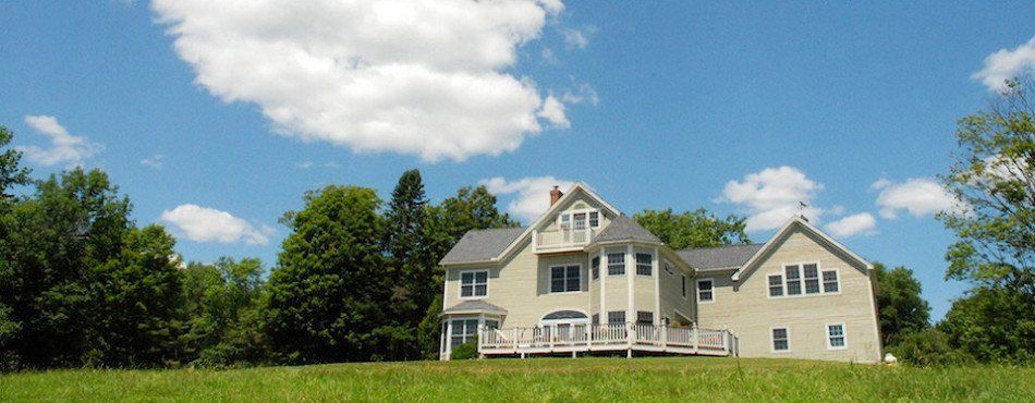 House with a light-colored exterior and gray roof set on a grassy hill with trees and a blue sky with white clouds.