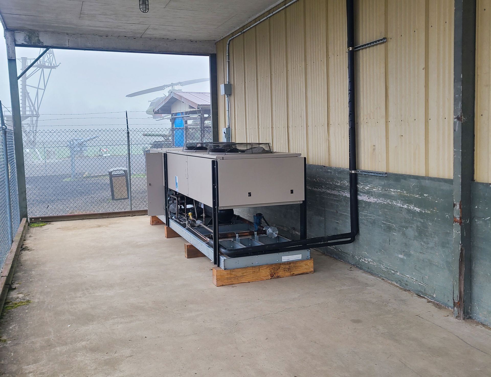 An industrial chiller unit on wooden blocks, mounted outside. Metal building with chain link fence visible.