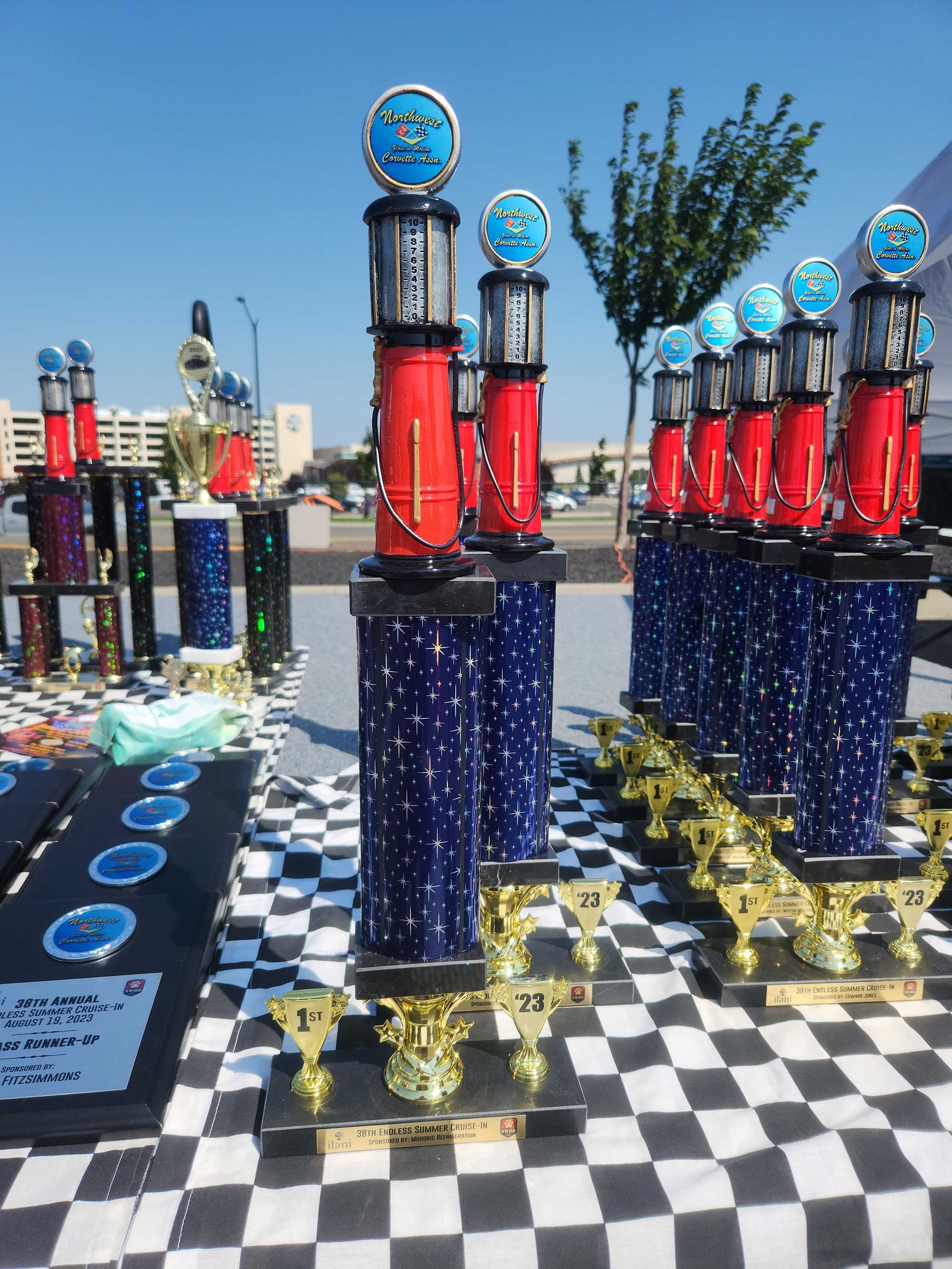 Trophies on a checkered table outside. Tall trophies are red and blue, shorter gold trophies in front. Bright, sunny day.
