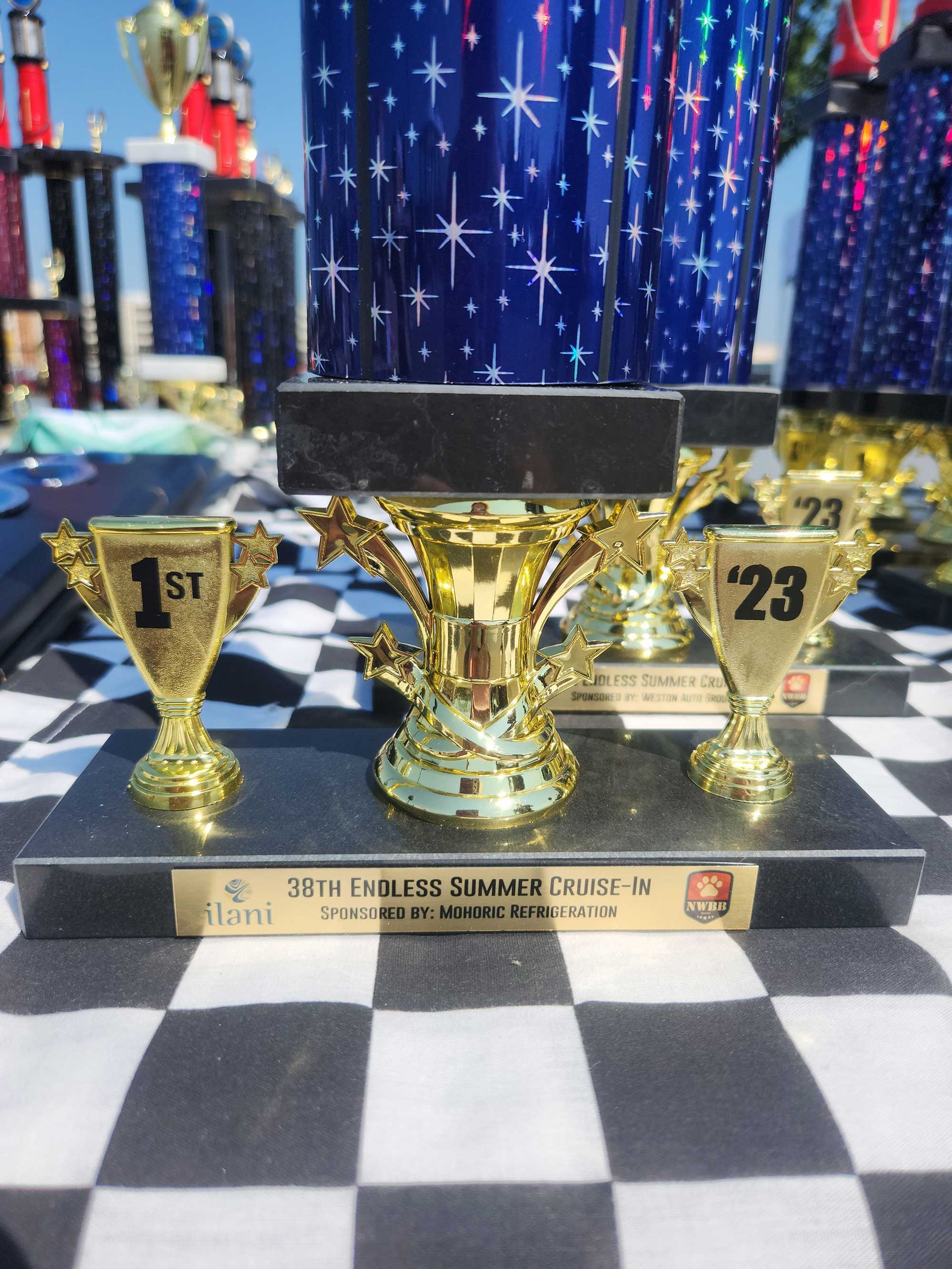 Trophies on a checkered surface, a large blue and gold trophy at center, smaller gold cups reading 