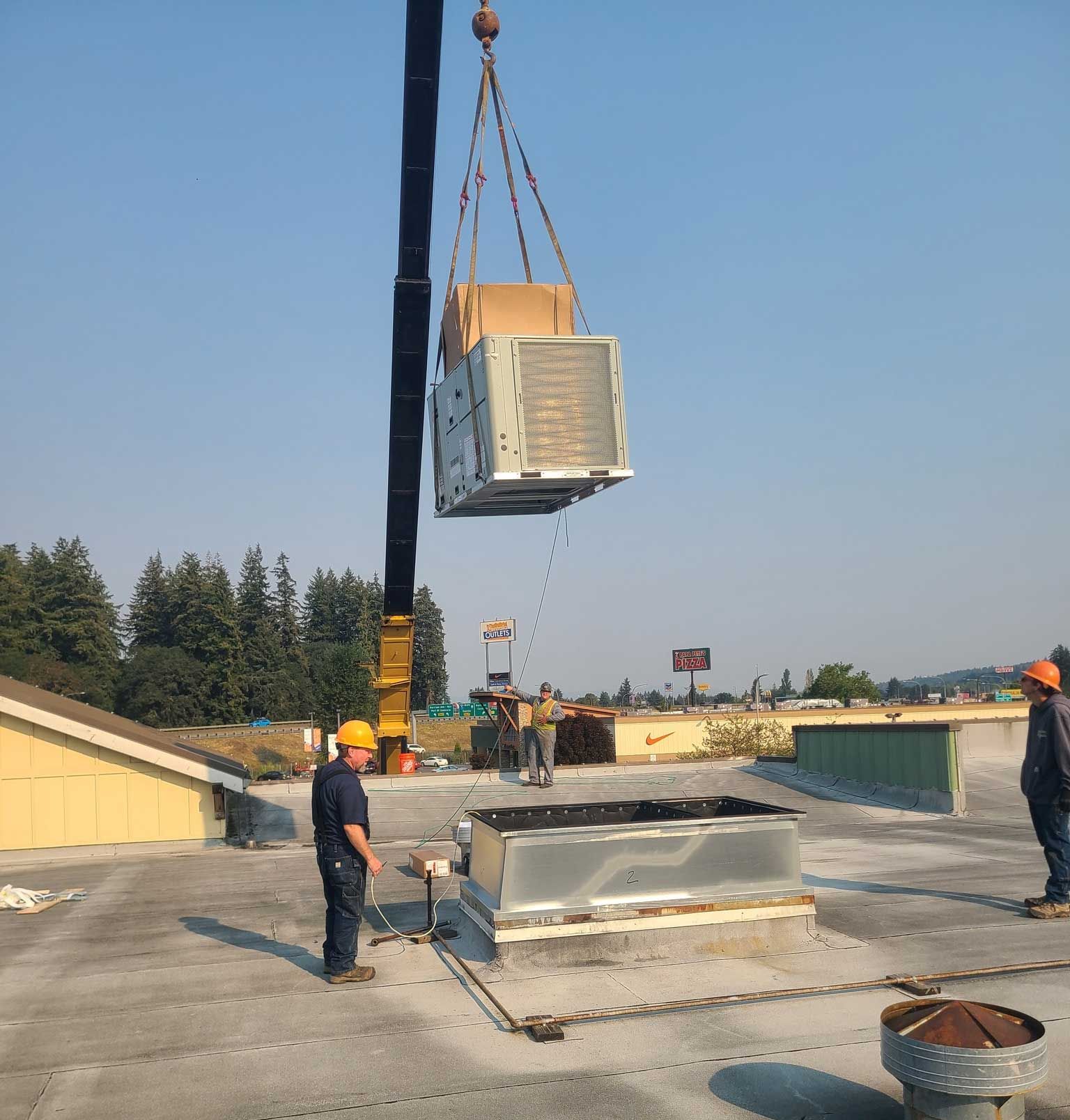 A crane lifts an HVAC unit onto a rooftop, as workers in hard hats observe.