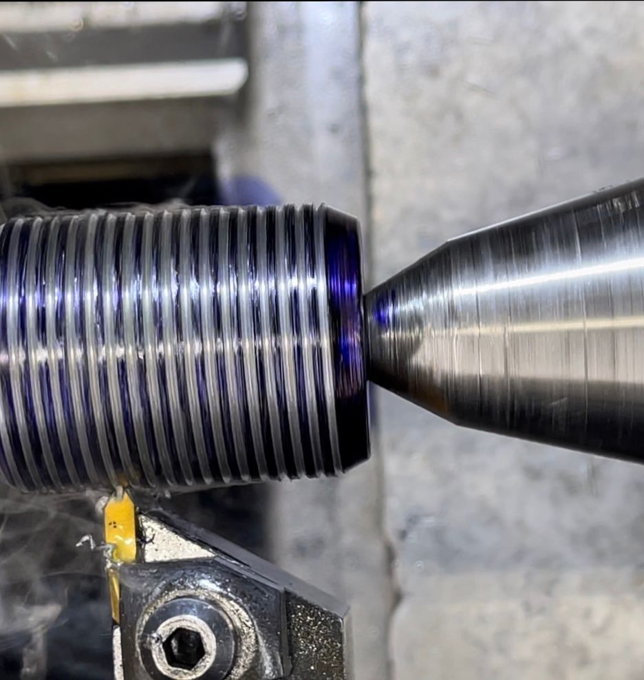 Close-up of a metal rod being threaded on a lathe with cutting tool. Purple metal shavings visible.