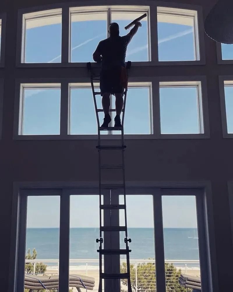 Worker on a ladder cleaning large windows with an ocean view behind them