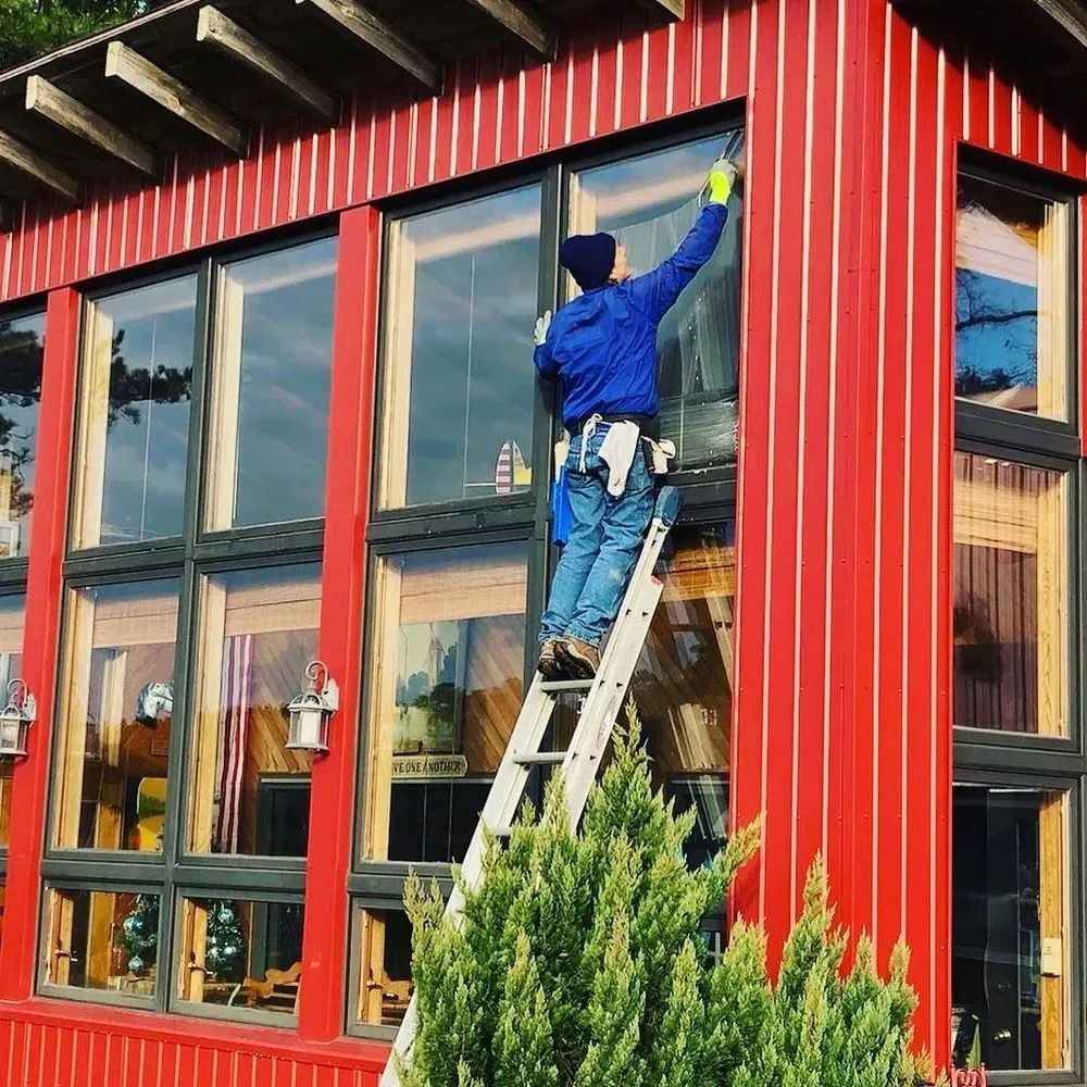 Worker on a ladder painting the red exterior trim of a building with large windows.