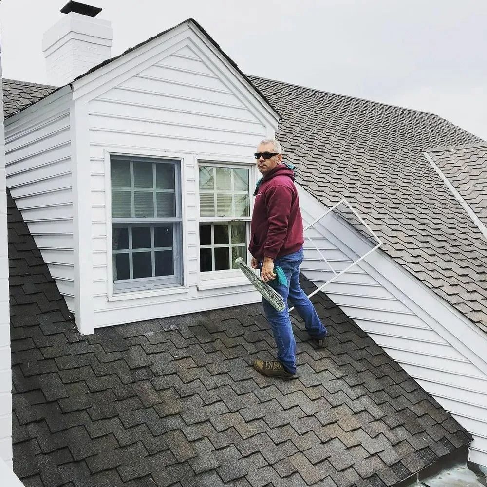 Worker standing on a steep shingled roof beside a white dormer window