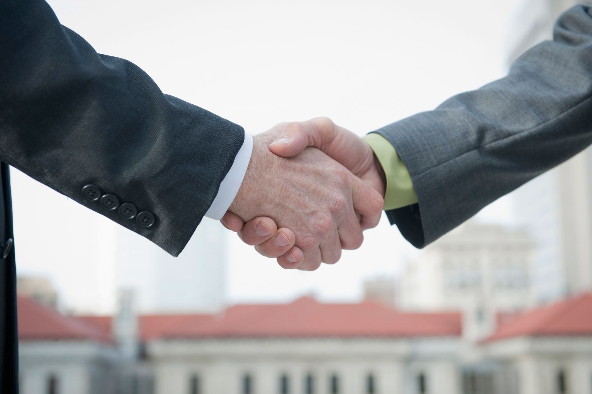 Two people in suits shaking hands, outdoors with building in background.