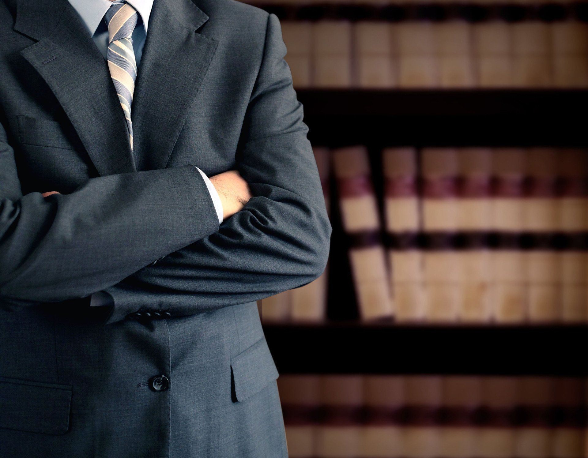 Man in suit with arms crossed, standing in front of a bookcase filled with books.